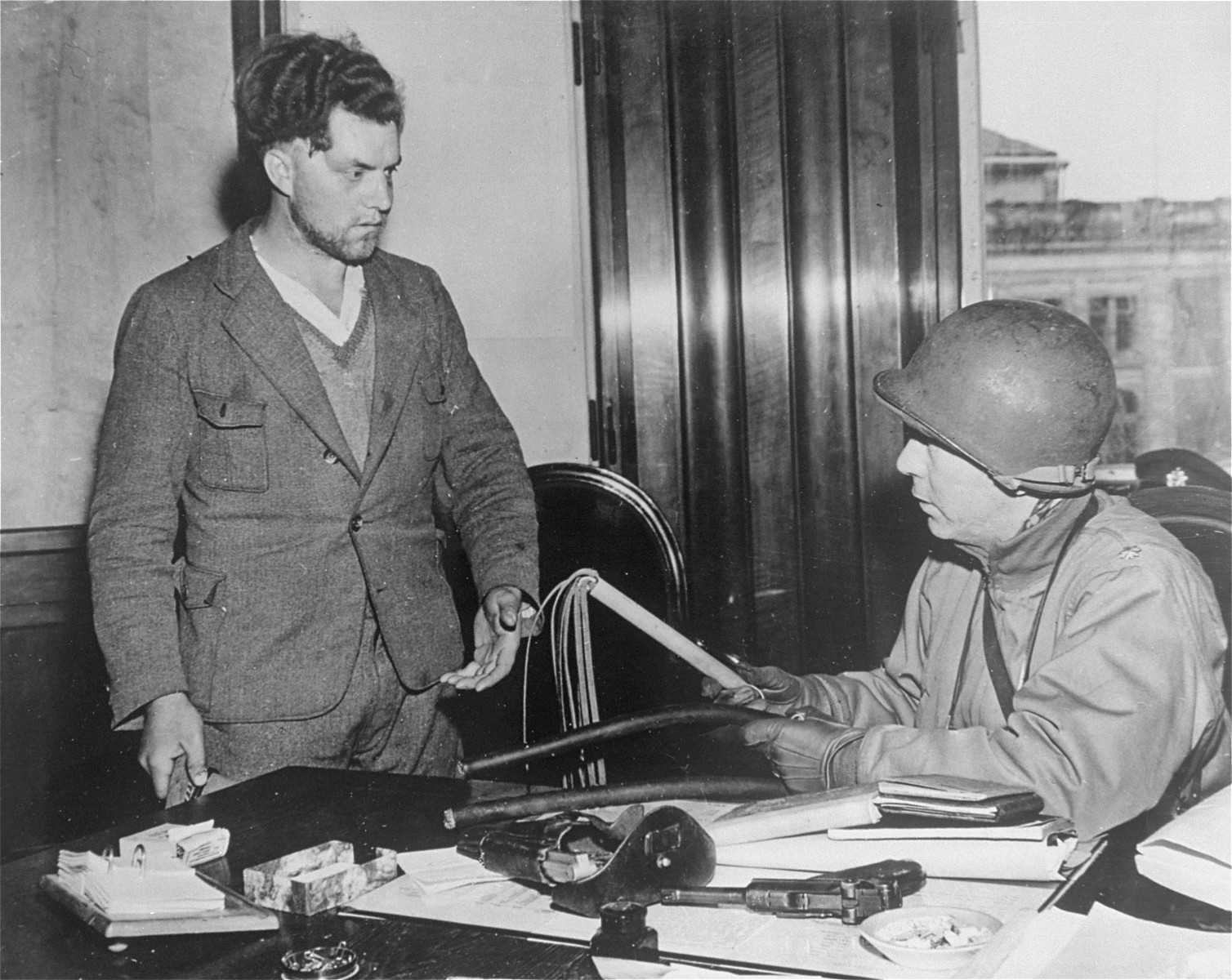 Major J.W. Bost of the 35th Infantry Division, Ninth U.S. Army, interrogates Lieutenant Herman Meyer (left), who was a Nazi storm trooper at the Lahde concentration camp near Hanover, Germany.  Bost is holding a leather thong whip which Meyer said he used only to "beat rugs."  Liberated prisoners claimed he used it to whip the prisoners.