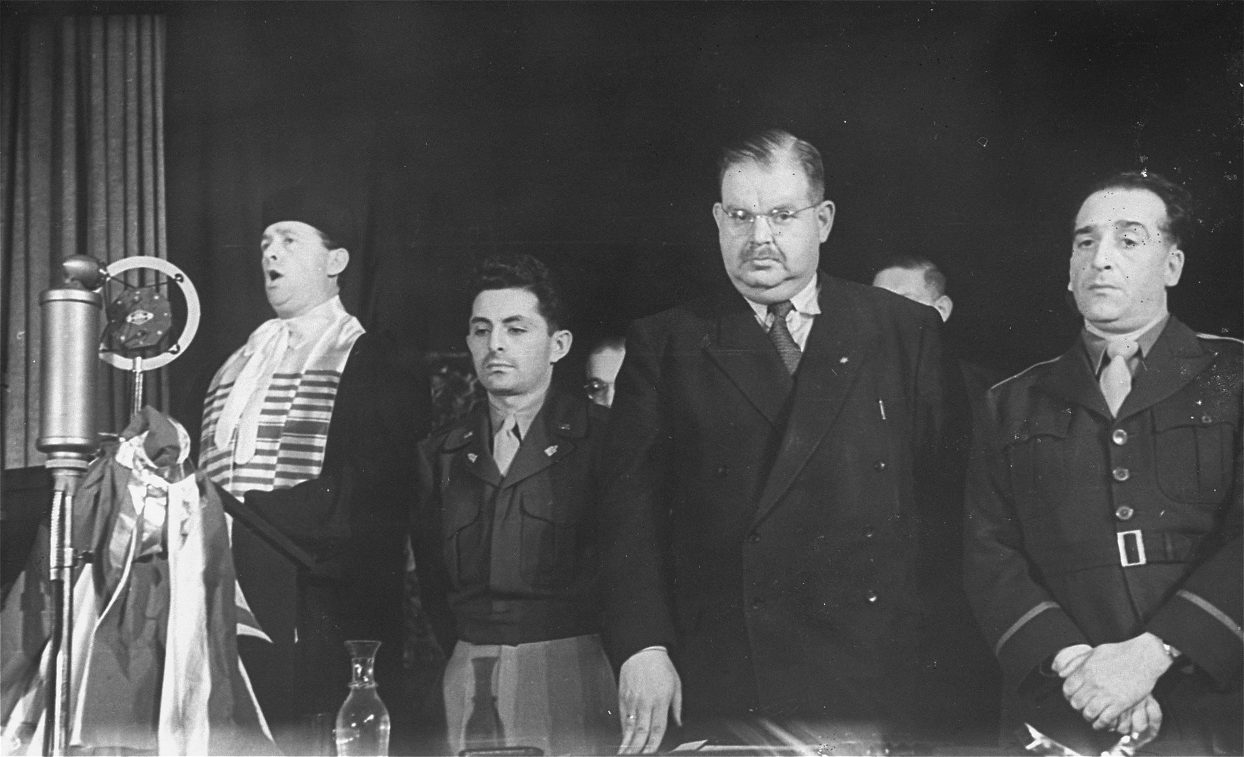 A cantor sings at a public ceremony sponsored by the Central Committee of Liberated Jews in the U.S. Zone of Germany.  

Among those pictured is U.S. Army chaplain Abraham Klausner (second from the left) and Philipp Auerbach (second from the right).