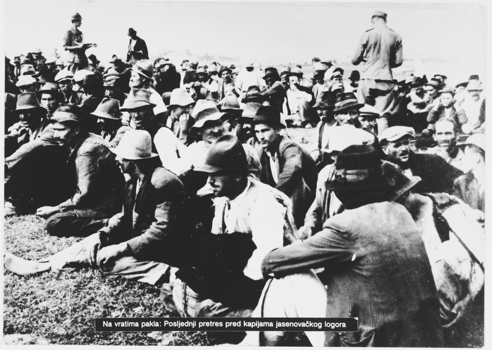 Ustasa guards move among a large group of Serbian villagers who are seated on the ground near the entrance to the Jasenovac concentration camp.

Original caption reads, "At Hell's door: last search at gates of camp Jasenovac"