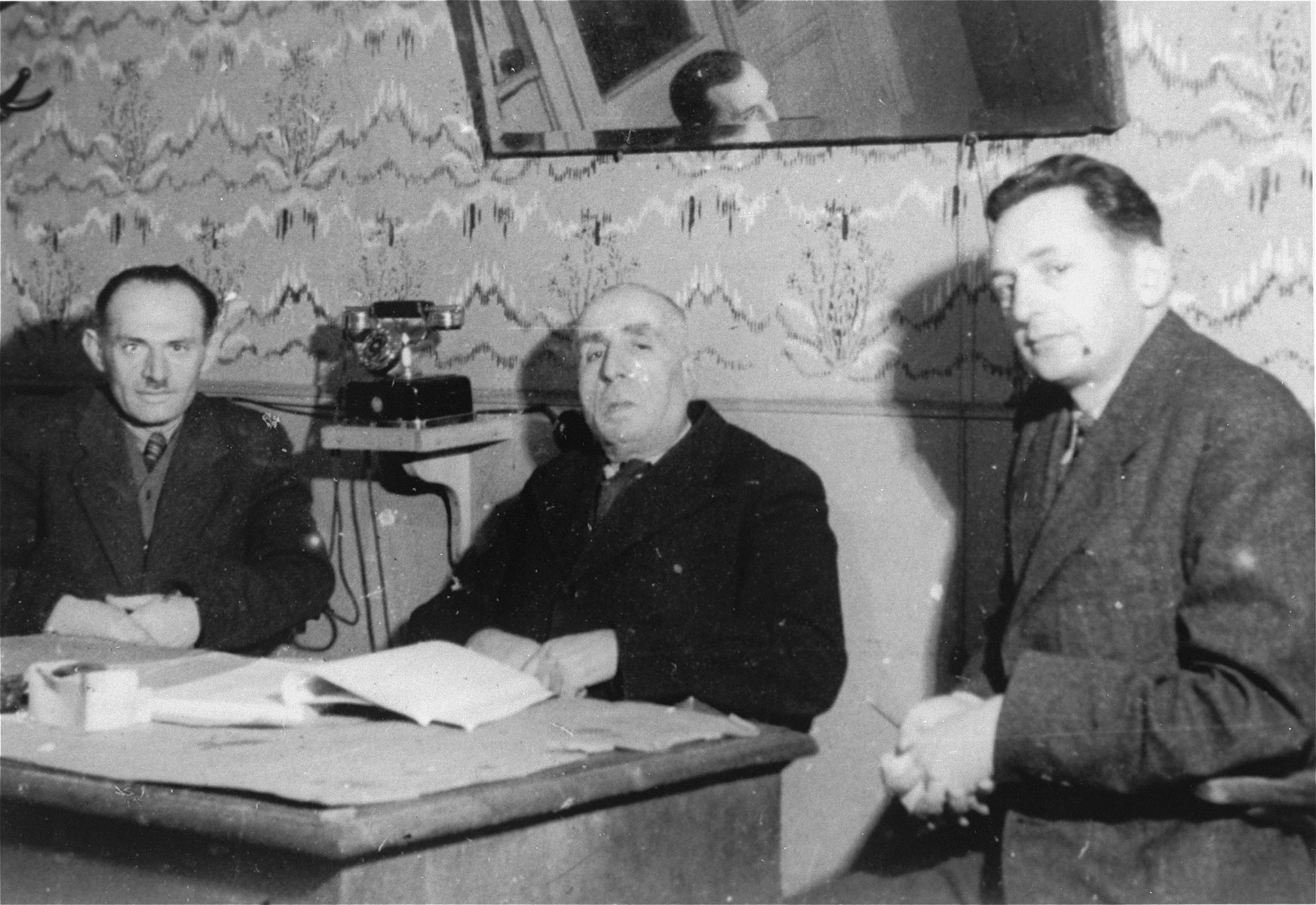 Three members of the ghetto administration sits around a table in the office of the Judenrat in the Kielce ghetto.

This photo was one the images included in an official album prepared by the Judenrat of the Kielce ghetto in 1942.