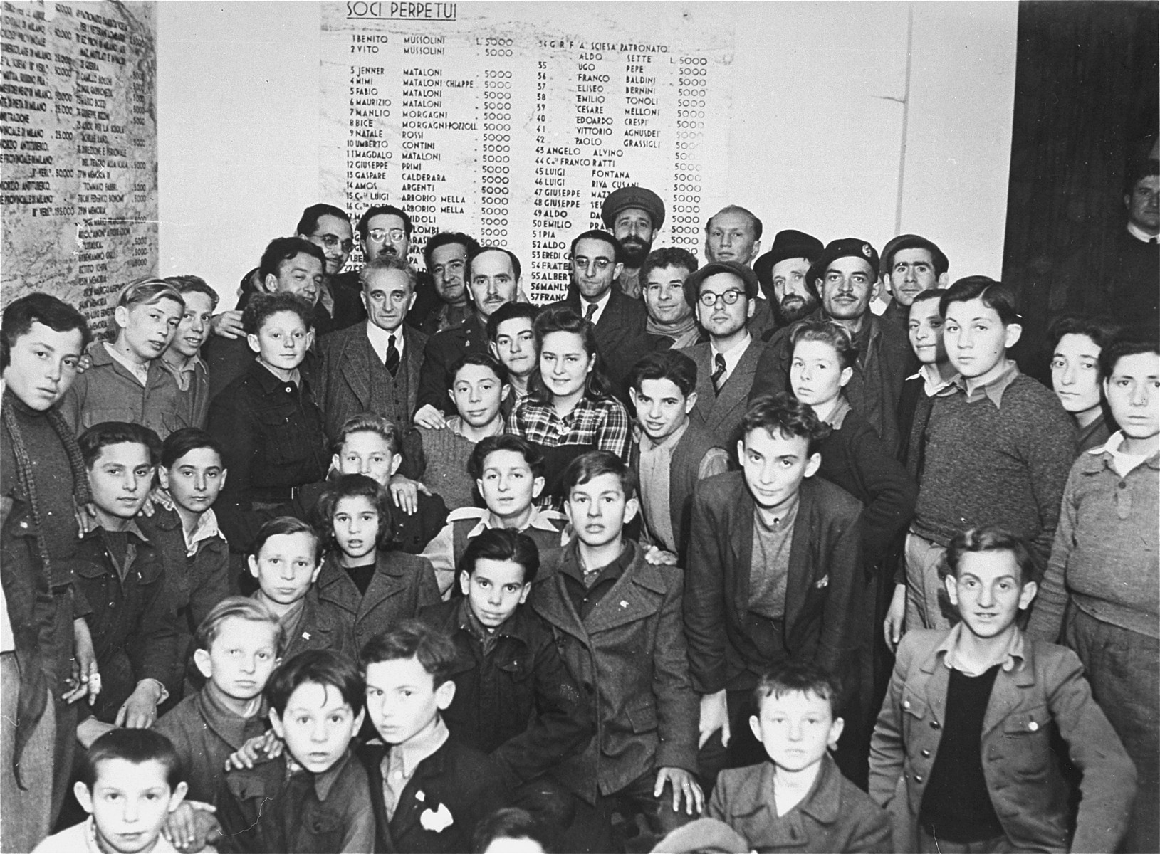 Group portrait of the staff and children of the Youth Aliyah children's home in Selvino, Italy.

The names on the wall are Italian donors who paid for Mussolini to build the villa as a Fascist youth center.

Among those pictured are Izak Abramowitz, Yaakov Meriash, Avraham Lipkunski, Jeshayahu Flamholz, Anita Teitelbaum, Moshe Blumenstock, Leon Lindenbaum, Matitiahu Drobles, Chaim Abramowitz, Surkis, Moshe Zeiri, Israel Drobles, Alpert, Szlama Fachter, and Morris (Moshe) Rubell.