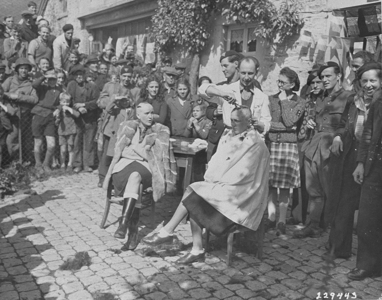 Belgian civilians and members of the resistance watch as a barber shaves the head of a woman who collaborated with the Nazis.  The woman at left has also had a swastika painted on her head.