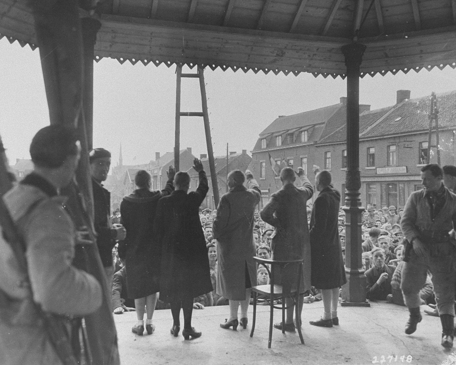 Belgian women who collaborated with the Germans during the occupation are forced to give the Nazi salute before their jeering countrymen.  The women's heads were all shaven as part of their public humiliation.