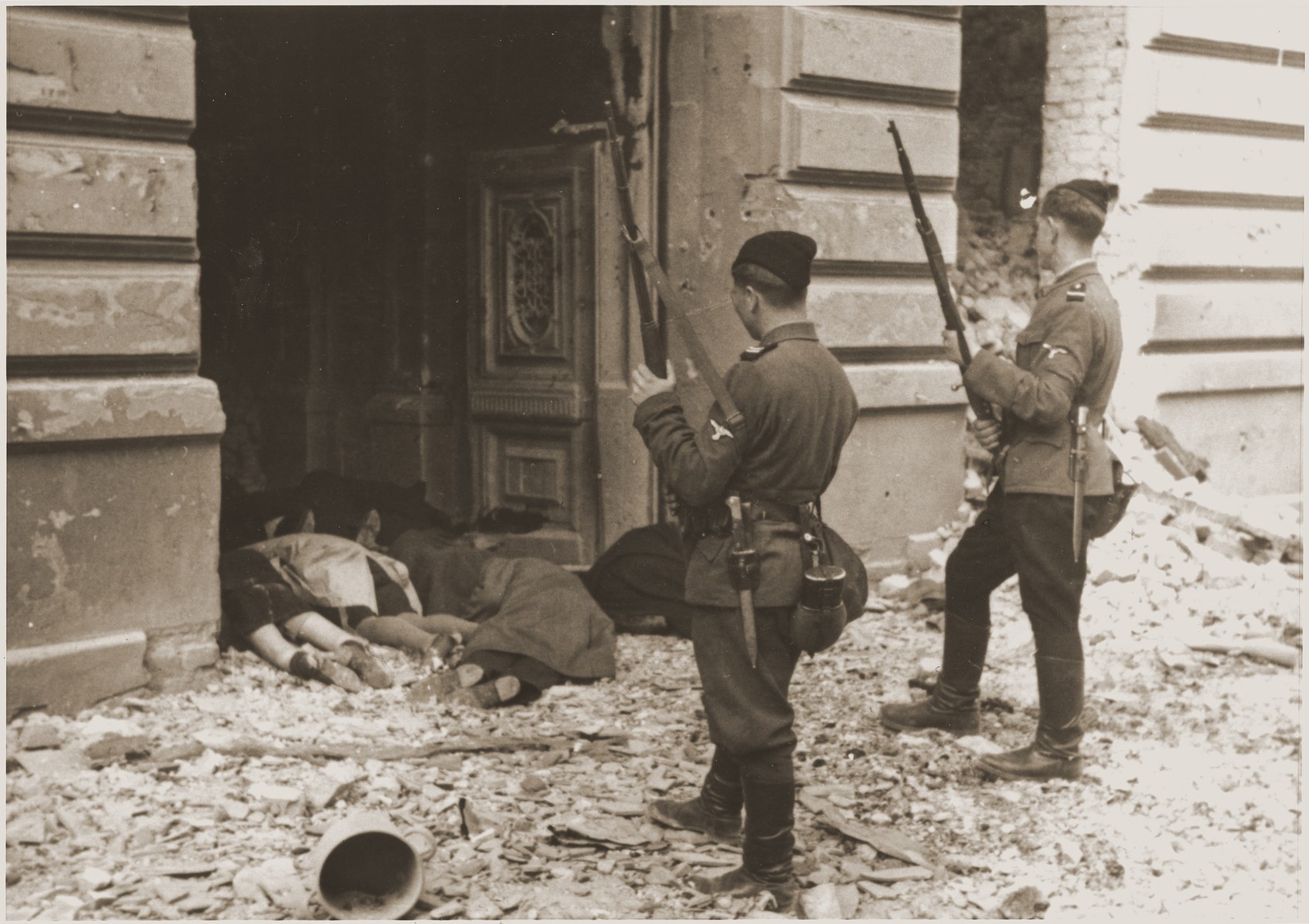 Askari or Trawniki guards peer into a doorway past the bodies of Jews killed during the suppression of the Warsaw ghetto uprising.  

The original German caption reads: "Askaris used during the operation."
