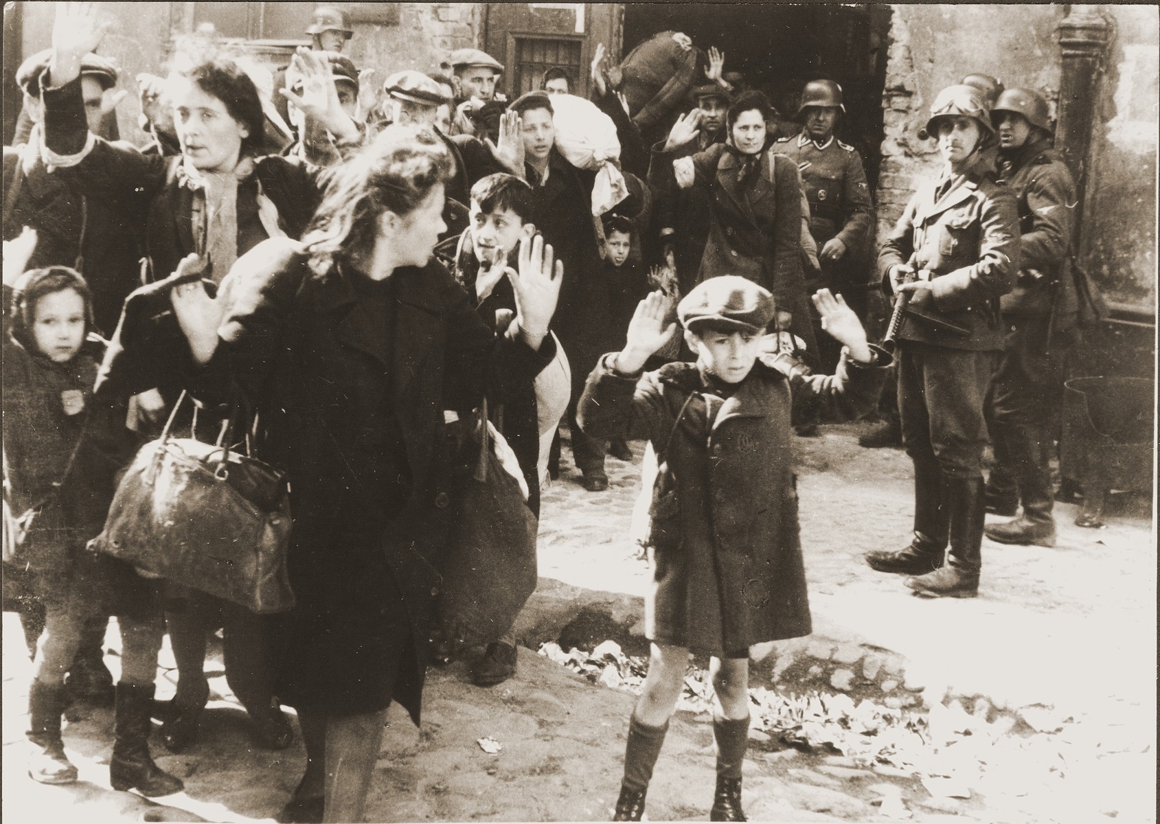 Jews captured by SS and SD troops during the suppression of the Warsaw ghetto uprising are forced to leave their shelter and march to the Umschlagplatz for deportation.  

The original German caption (translated to English) reads, "Pulled from the bunkers by force."

The SD trooper pictured second from the right, is SS-Rottenfuehrer Josef Bloesche, who was identified by authorities using this photograph.  Bloesche was tried for war crimes by an East German court in 1969, sentenced to death and executed in July of that year.  

The little girl on the left has been identified as Hanka Lamet, who is standing next to her mother, Matylda Lamet Goldfinger (the woman second from the left).  The boy carrying the sack has been identified as Leo Kartuzinsky and the woman in the front has been identified as Chana Zeilinwarger.  Numerous people have identified the boy in the foreground as either Arthur Domb Semiontek, Israel Rondel, Tsvi Nussbaum or Levi Zeilinwarger, but none of these identifications can be conclusively corroborated.

Original caption from donated photograph reads:  "The following pictures are copies of the book containing the report of the German commander who was responsible for cleaning out that ghetto in Warsaw, Poland.  This report, in the form of a leather bound book, was presented as evidence by the U.S. prosecution, Maj. Frank Walsh, at the international tribunal trials at Nurnberg, Germany."