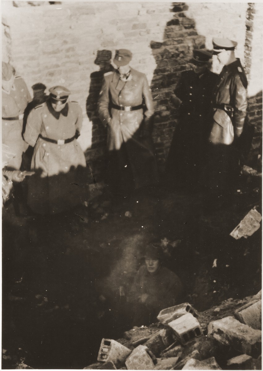 SS Major General Juergen Stroop (center), accompanied by SS and police officers, looks on as SS soldiers uncover the entrance to an underground bunker during the suppression of the Warsaw ghetto uprising.  The original German caption reads,  "They were also discovered in underground bunkers."