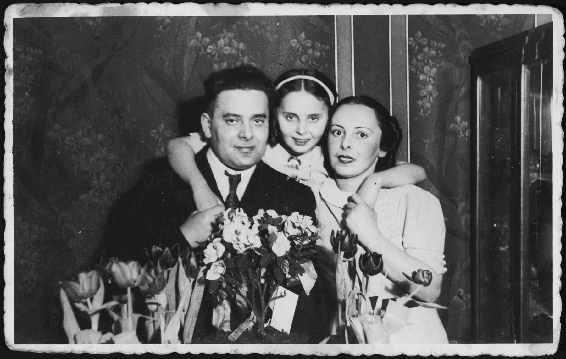 A young girl poses between her parents surrounded by flowers during their 10th anniversary party.

Pictured are Josef, Rina and Ada Altbeker.