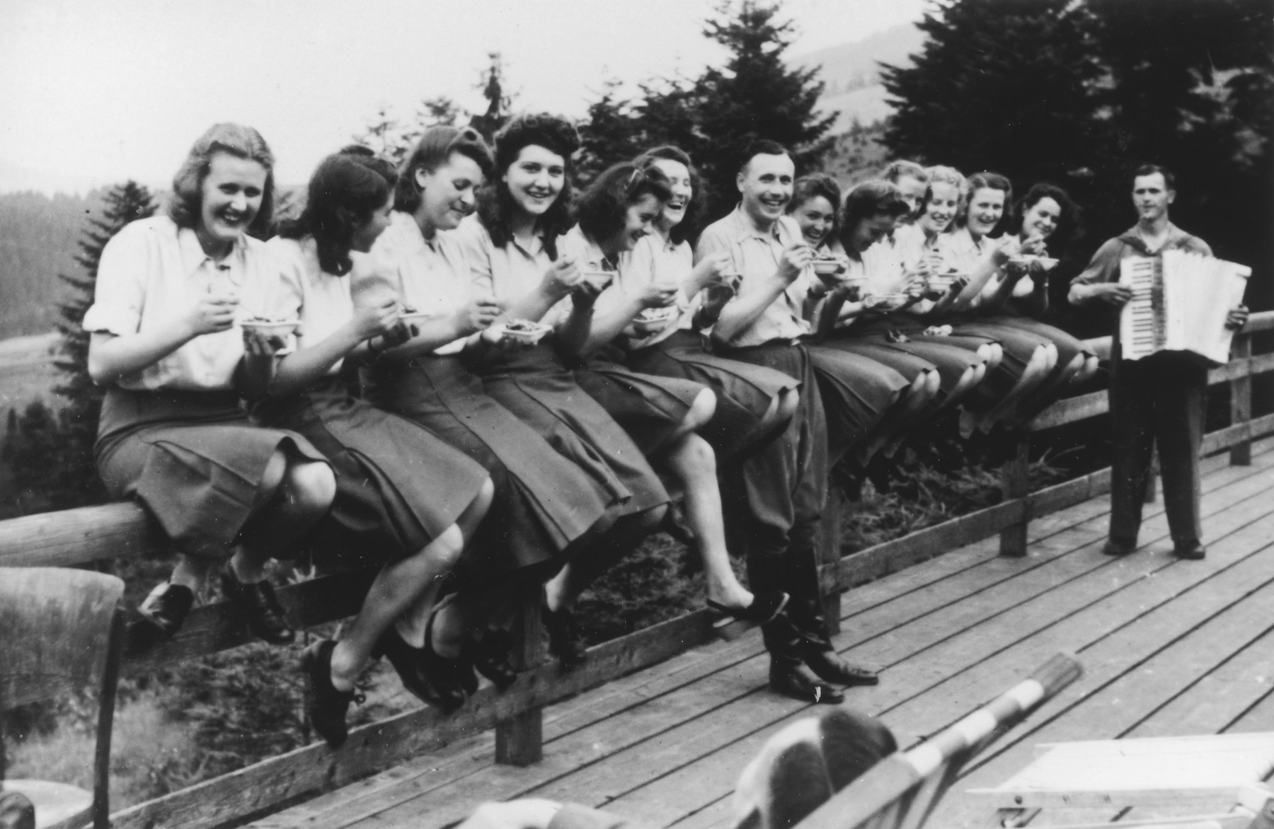 Members of the SS Helferinnen (female auxiliaries) and SS officer Karl Hoecker sit on a fence railing in Solahuette eating bowls of blueberries.  In the background is a man playing the accordion. 

The original caption reads "Hier gibt es Blaubeeren" (there are blueberries here).