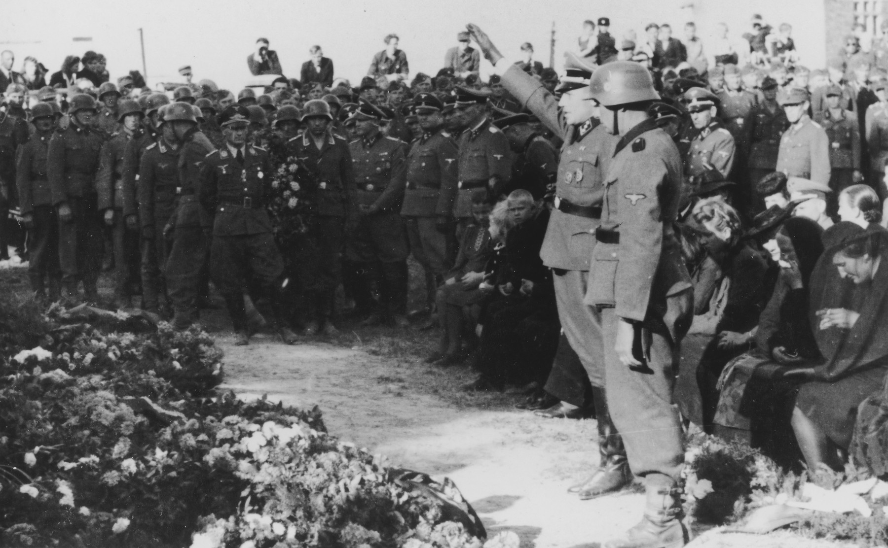 SS officer, Karl Hoecker salutes in front of an array of wreaths during a military funeral near Auschwitz.

The original caption reads "Beisetzung von SS Kameraden nach einem Terrorangriff."  (Burying our SS comrades from a terror attack.) 

This probably is the aftermath of the September 13th bombing of IG Farben in which 15 SS men died in the SS residential blocks and 28 were seriously wounded.

Pictured in the background are Josef Kramer and Karl Moeckel.