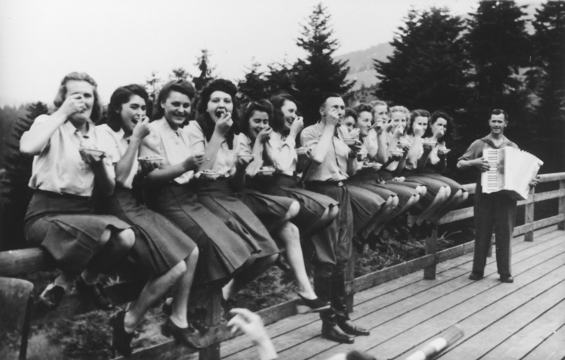 Members of the SS Helferinnen (female auxiliaries) and SS officer Karl Hoecker sit on a fence railing in Solahuette eating bowls of blueberries.  In the background is a man playing the accordion. 

The original caption reads "Hier gibt es Blaubeeren" (there are blueberries here).