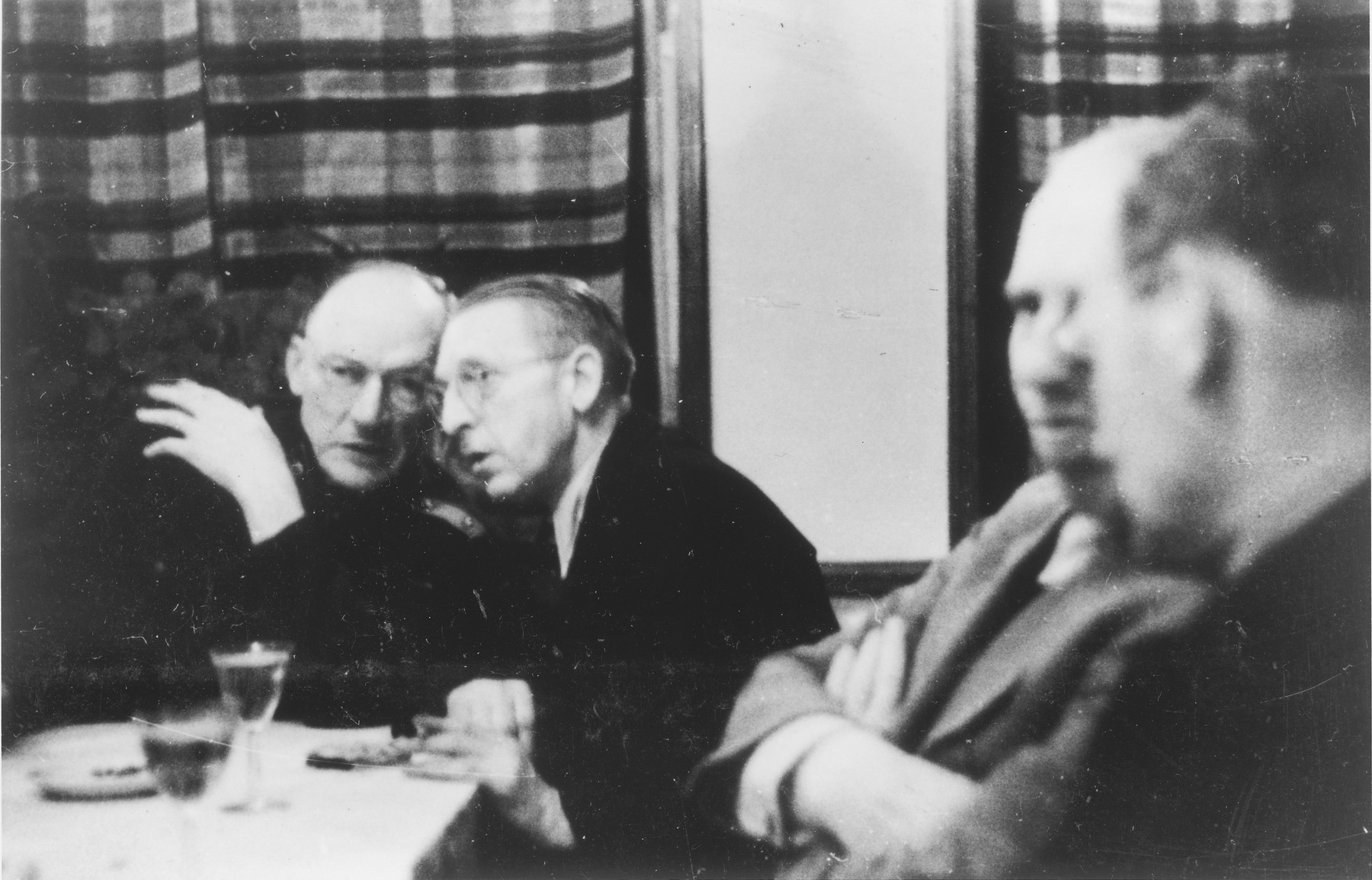 Four Germans chat amongst themselves around a table in a hunting lodge. 

Heinrich Josten is pictured on the left.