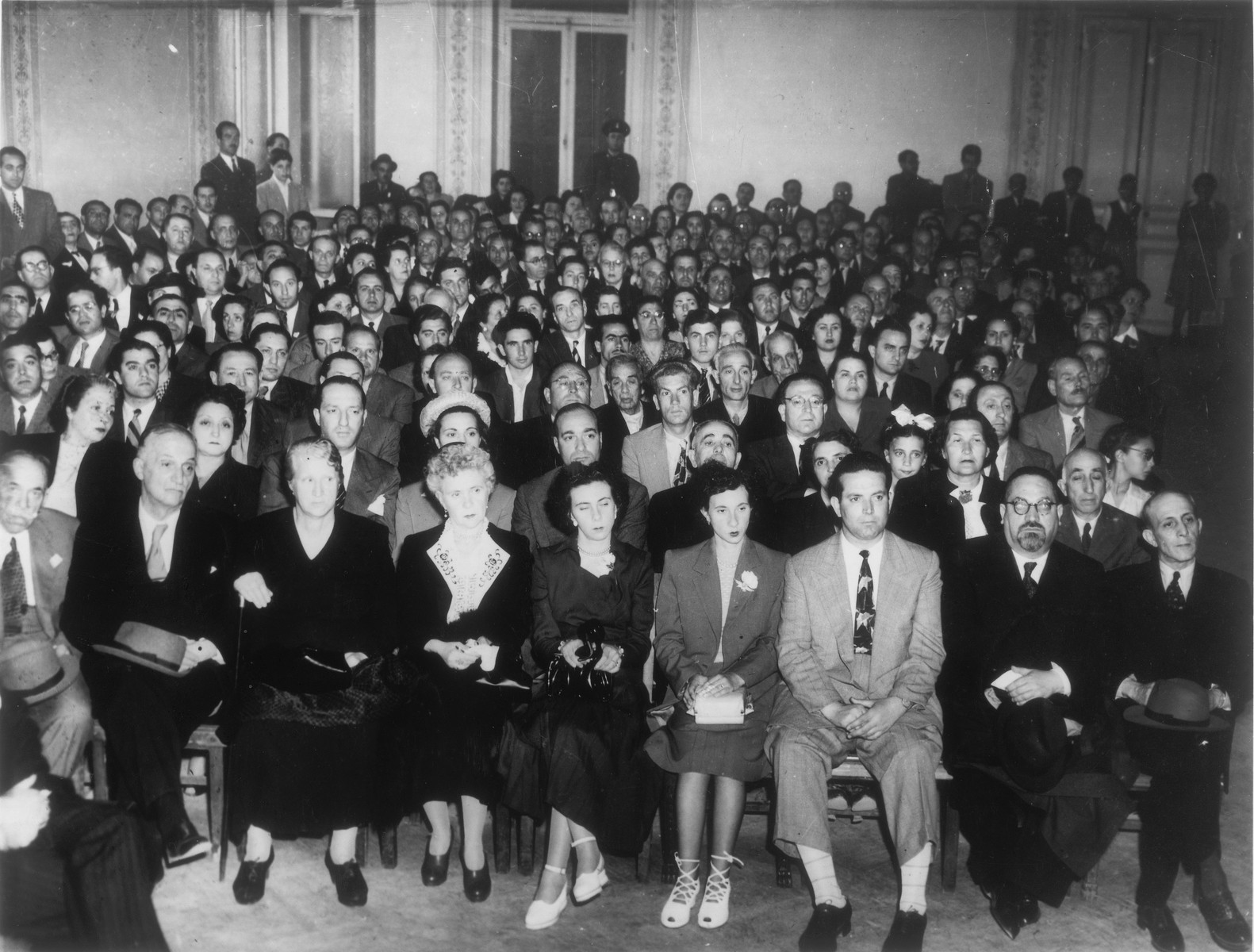 A large crowd assembles in the Parnassos lecture hall in Athens to celebrate the founding of Israel.  

Rabbi Elias Barzilai is seated in the front row, second from the right.  Jack Amar is seated on the far right.  Also pictured are Noah Yeshouroun, Sylvia Levy, Freddy Barouh, Rosetta Vital, Noulis Vital, Elias Leon, Markos Tabach, Michael Matsas, Pepo Benouziglio, Lydia Shabetay, and Kaimis.