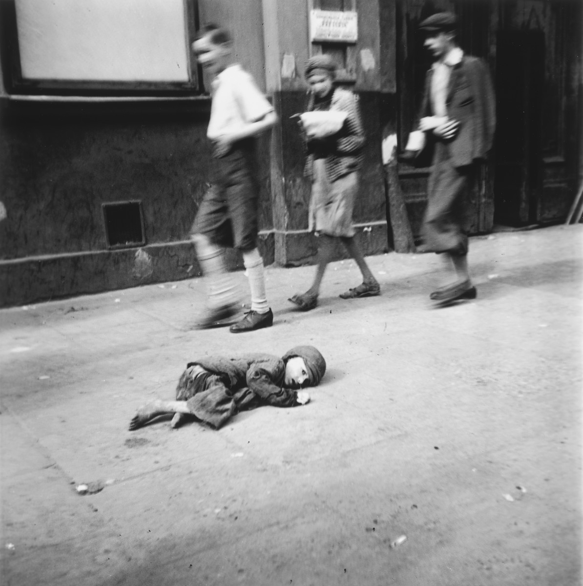 A destitute young boy lies on the pavement in the Warsaw ghetto as other children walk by.

Joest's original caption reads: "On a sidewalk in a side street I saw this tiny child who could no longer pull himself upright.  The passers-by didn't stop.  There were too many children like this one."