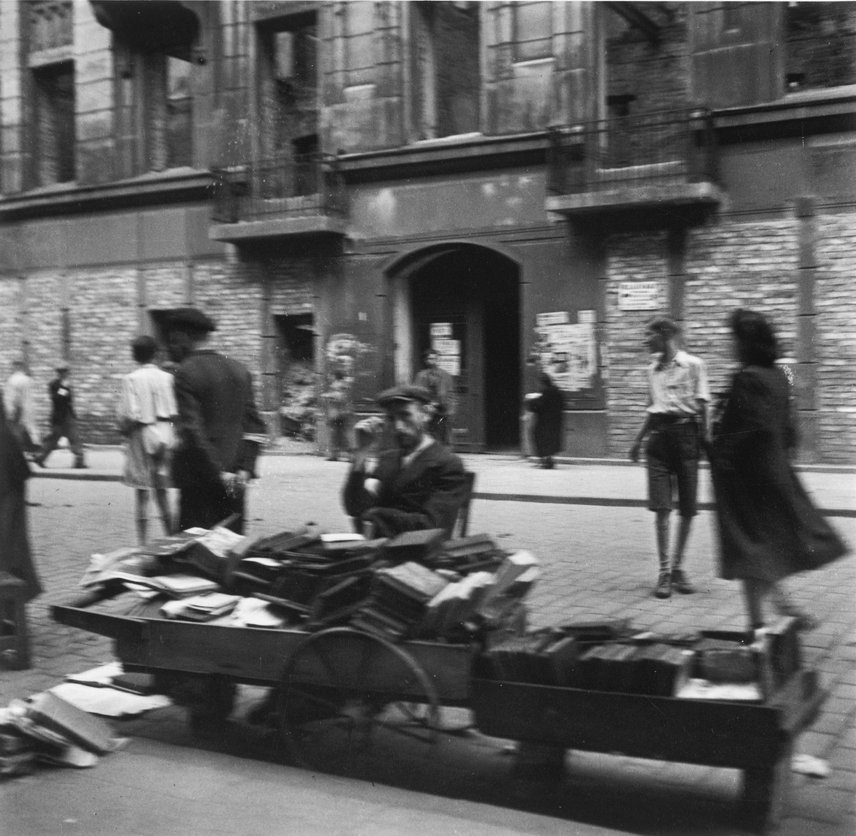 A vendor sells used books on a street in the Warsaw ghetto.

Joest's original caption reads: "I remember quite clearly that I asked myself how these people could buy books when they had nothing to eat.  I don't recall the location of this picture.  There were many such book carts."