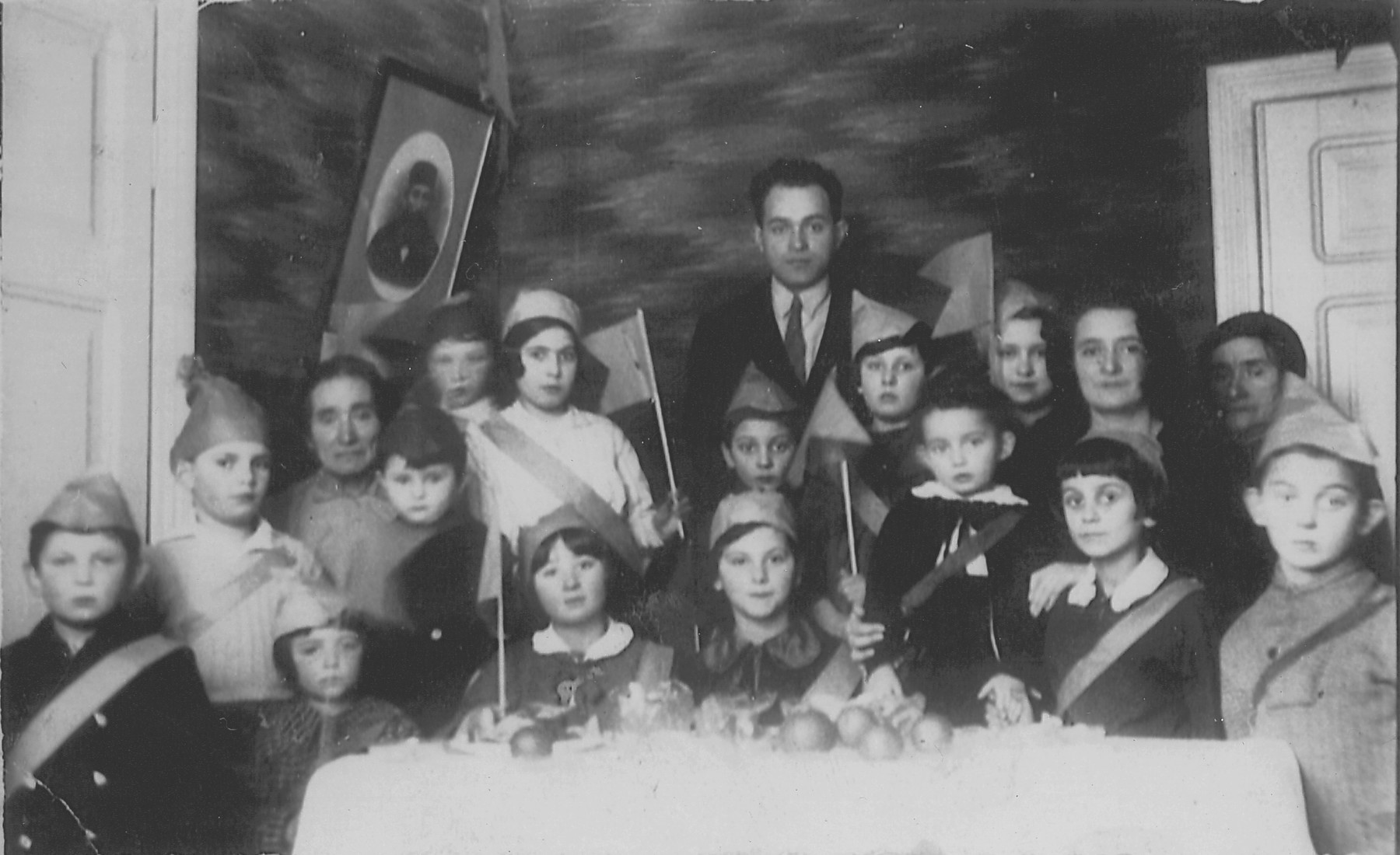 Lejb Melamdowicz's third birthday, Bialystok, 1935. 

Melamed's father (center), grandmother and mother (far left and far right, respectively) join a group of children in hats and sashes around a table adorned with fruit.