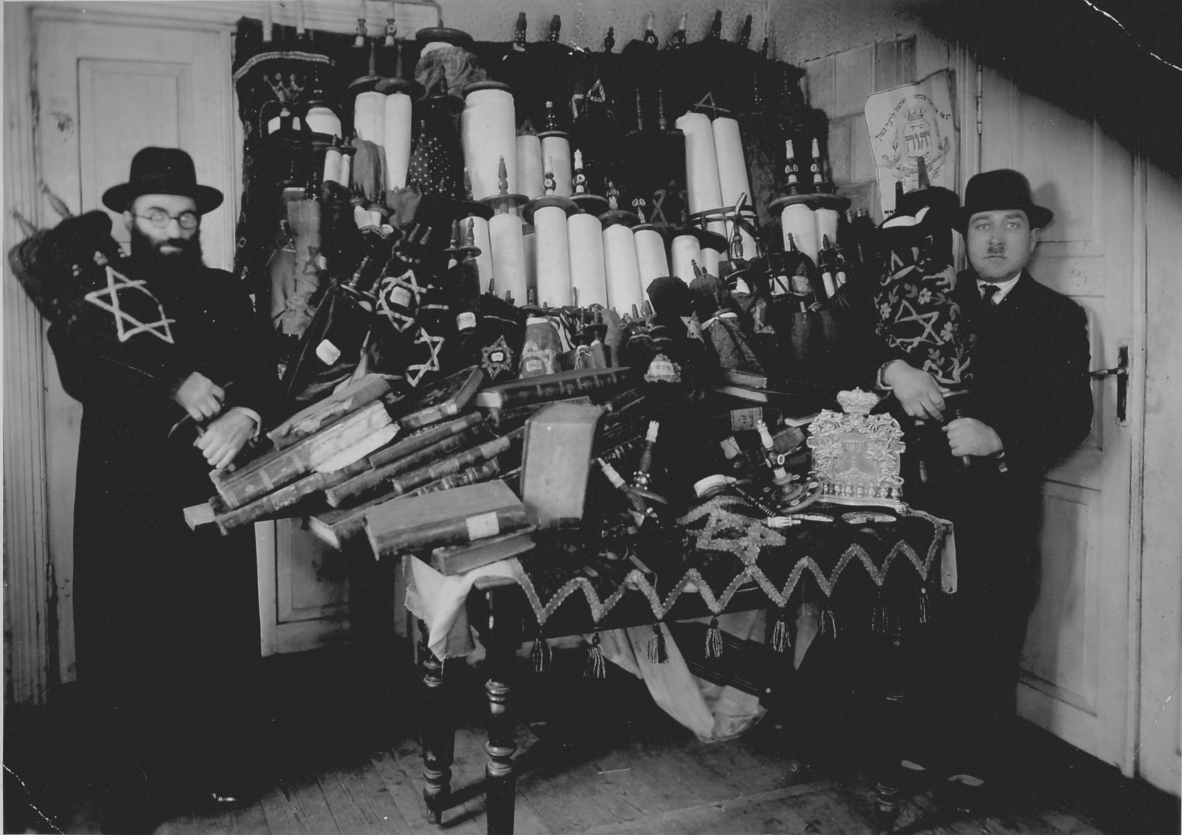 Rabbi Lifszyc (left) and another man pose in front of a cabinet full of Torah scrolls that were smuggled out of Suwalki.

Sixty scrolls and other holy items were rescued from hiding places in Suwalki, which had been occupied by Germany, and were taken to the synagogue in Kalvarija for safety.  While Lifszyc was in Vilnius, he tried to save the Torah scrolls from the Russian occupiers, but had to flee Vilnius and leave these behind.