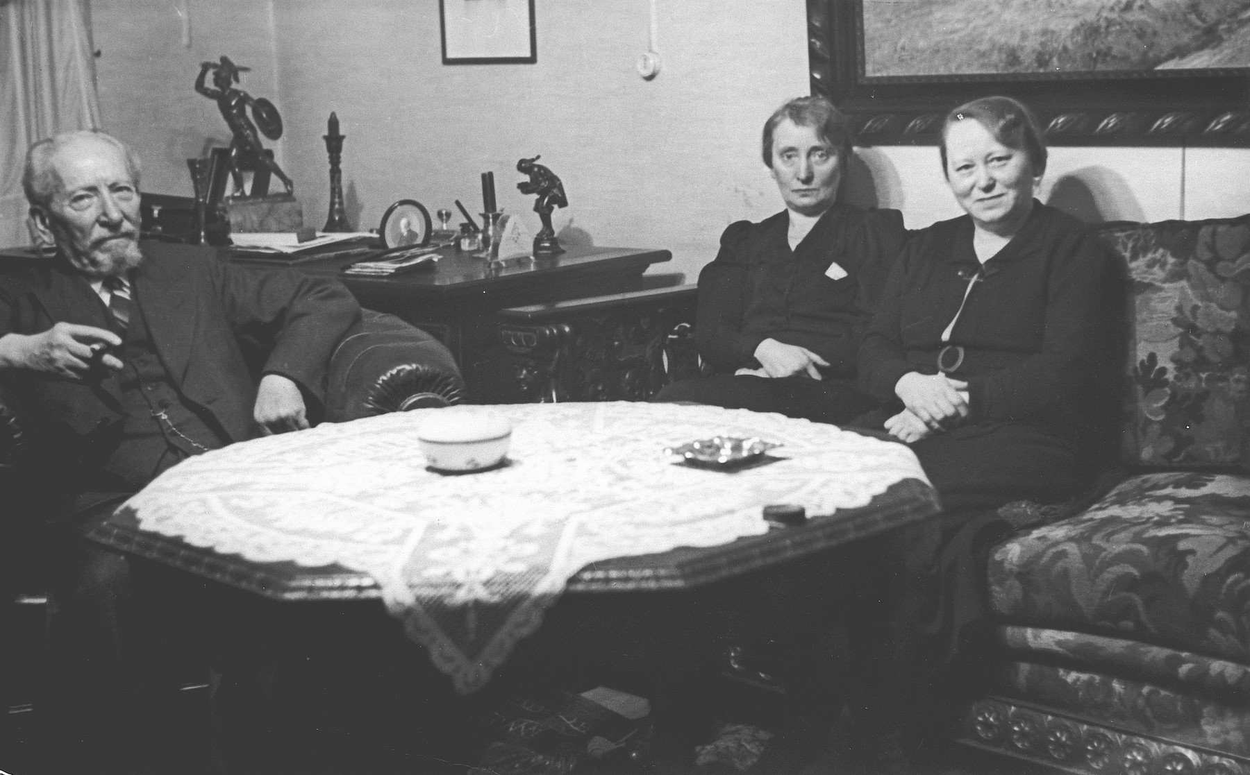 A German-Jewish family sits around a table in their home in Kassel.  

From left to right are Heinrich Bluth and his daughters Rosa and Regina.