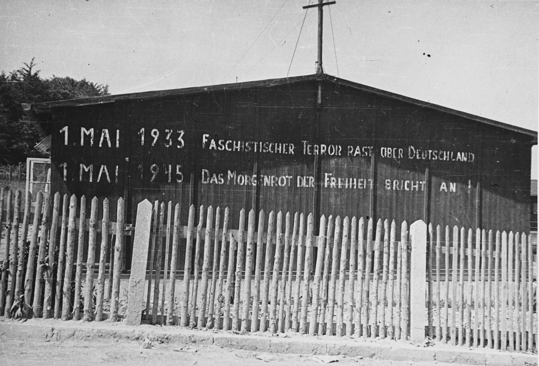 View of an inscription printed in large block letters on the wall of a barrack in the Buchenwald concentration camp.

The inscription on the building reads, "1 May, 1933  Fascist terror reigns over Germany.  1 May, 1945 Sunrise brings the dawn of freedom."

The original photo caption reads, "Shows where people have been there since 1933.  Buchenwald was in full swing about 1935.  Thousands died daily from typhus, dysentery, and T.B.  Most of them died in the crematory or the lime pit."