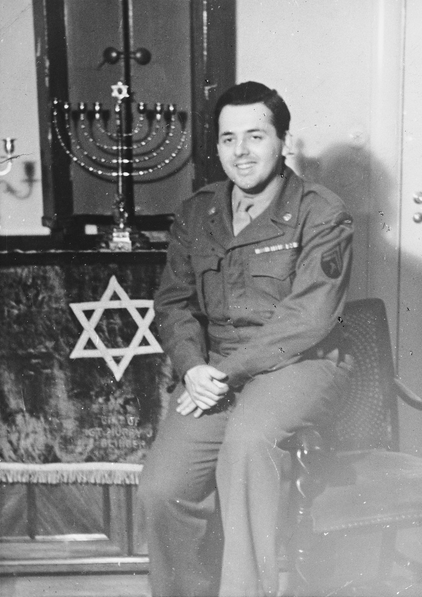 Close-up portrait of Saul Loeb, a Jewish chaplain's assistant, sitting in front of the Torah ark and menorah in the Berlin Chaplain's center.

The original caption reads, "Another picture taken in front of our Ark -- the ark was made especially for Chaplain Shubow by a Jewish architect from Holland, and he carried it in his jeep for services at the front during combat."