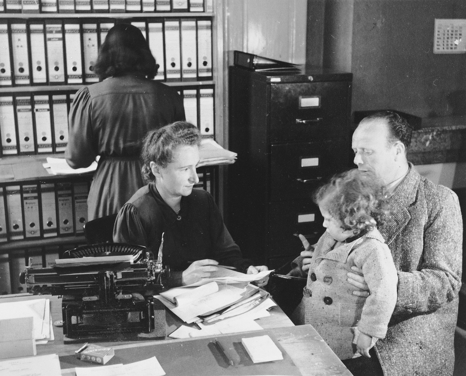 A woman, herself a refugee from France, assists a gentleman with a young child on his lap, in the offices of Secours aux Refugies in Geneva, Switzerland.

Seated at the desk is Ines Limot.