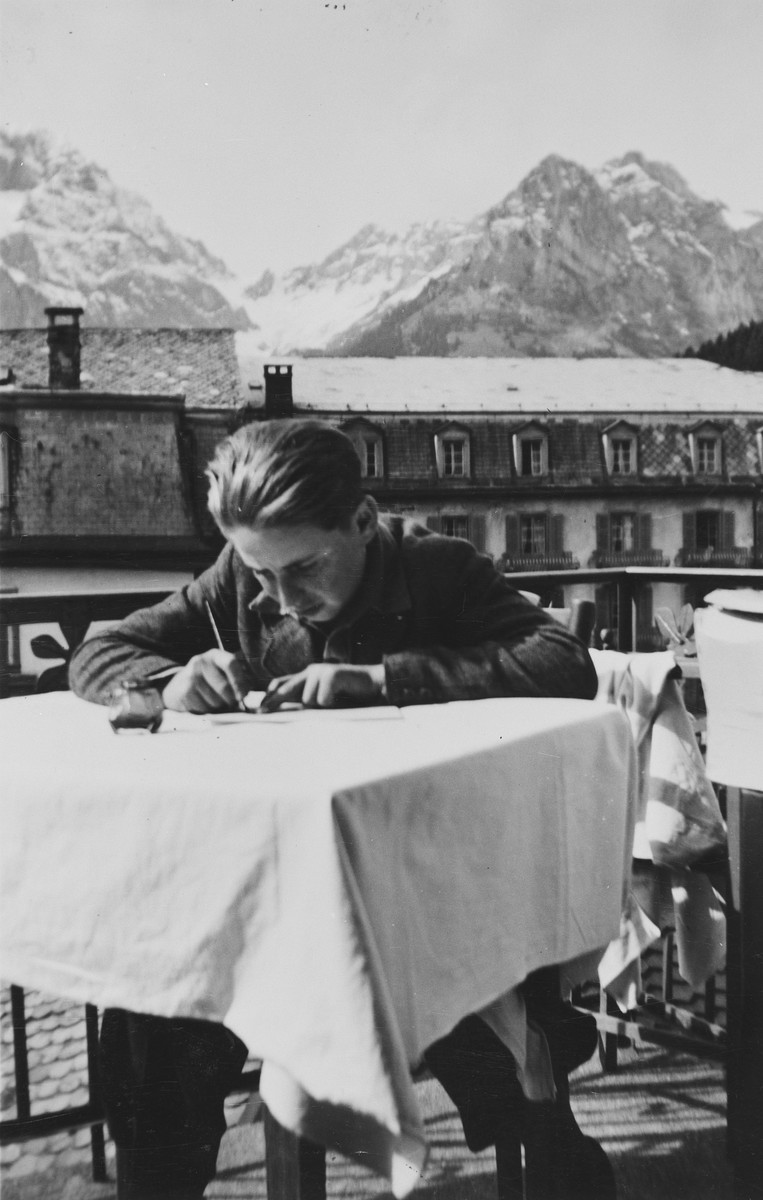A young Jewish man writes at a table in a children's home in Switzerland.  The snow capped Alps can be seen in the background.