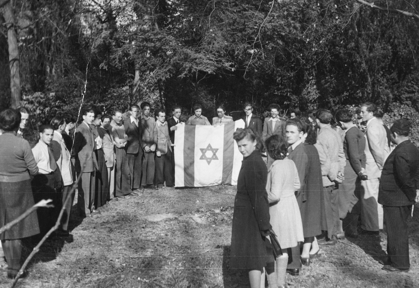 Jewish youth gather for a ceremony around a large Zionist flag in a children's home in Switzerland.

Moniek Szmulewicz is among those pictured.  Monek 
Goldfinger is standing on the far right in a light colored suit.