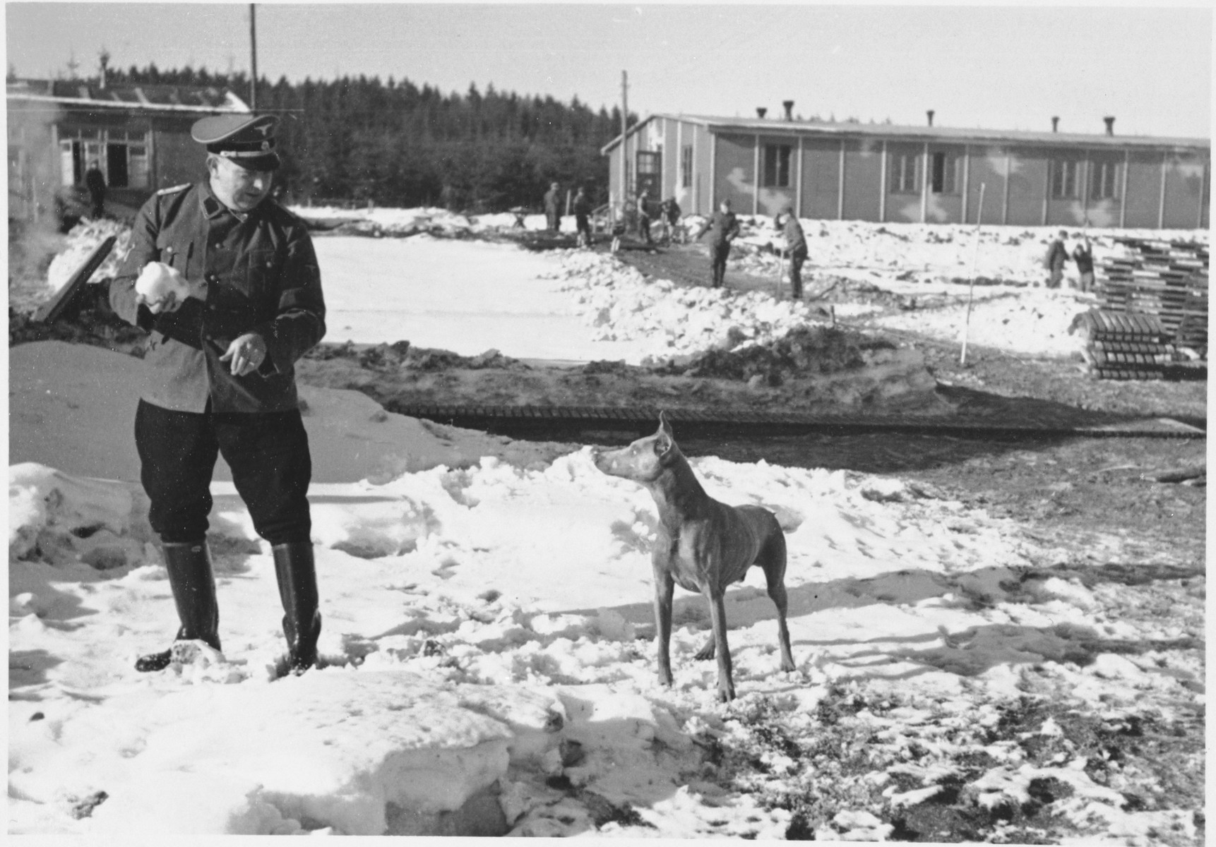 A Nazi official plays with his dog in the snow at Hinzert (sub-camp of Buchenwald)  while prisoners can be seen working in the background.