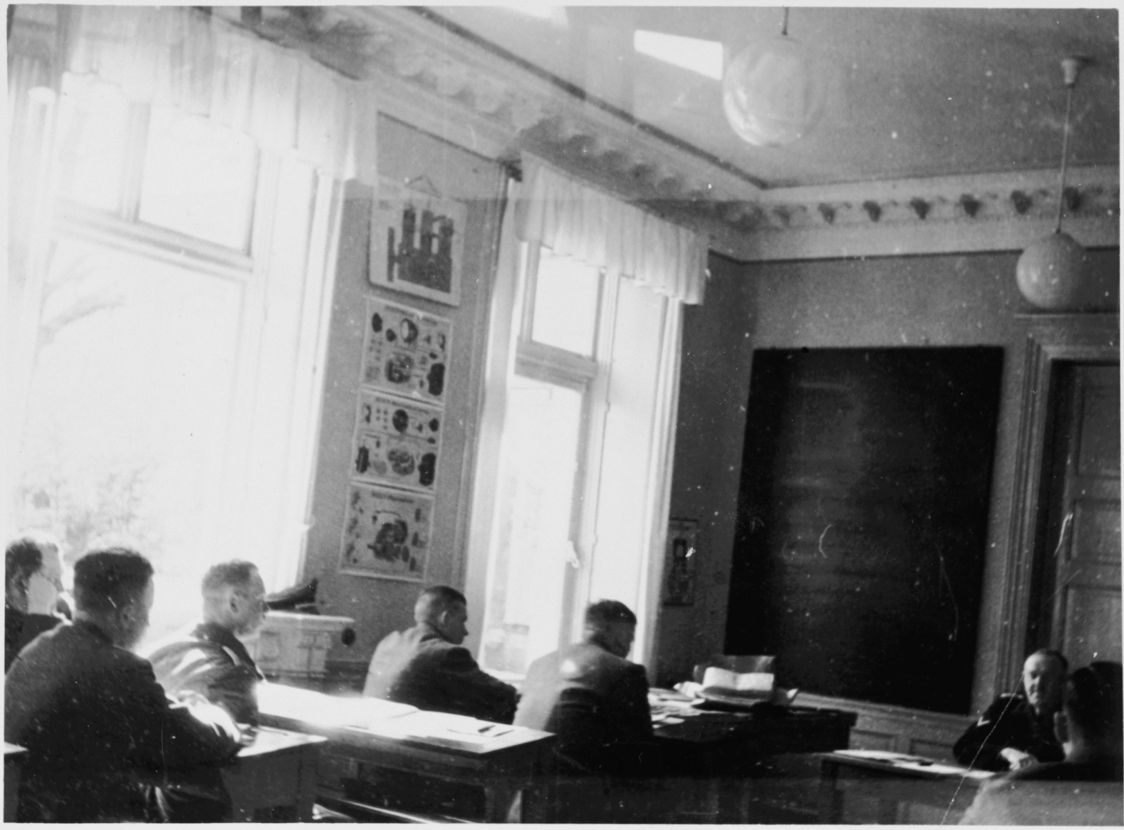 SS officers sit in a classroom in the Hinzert concentration camp (a sub-camp of Buchenwald).

The classroom has been tentatively identified as the "Schulungsraum"  (training room) and the instructor as Prof. Dr. Gustav Riek.  The photograph may have been taken May 7, 1941, during the inspection visit of Adolf Kleffel and others.