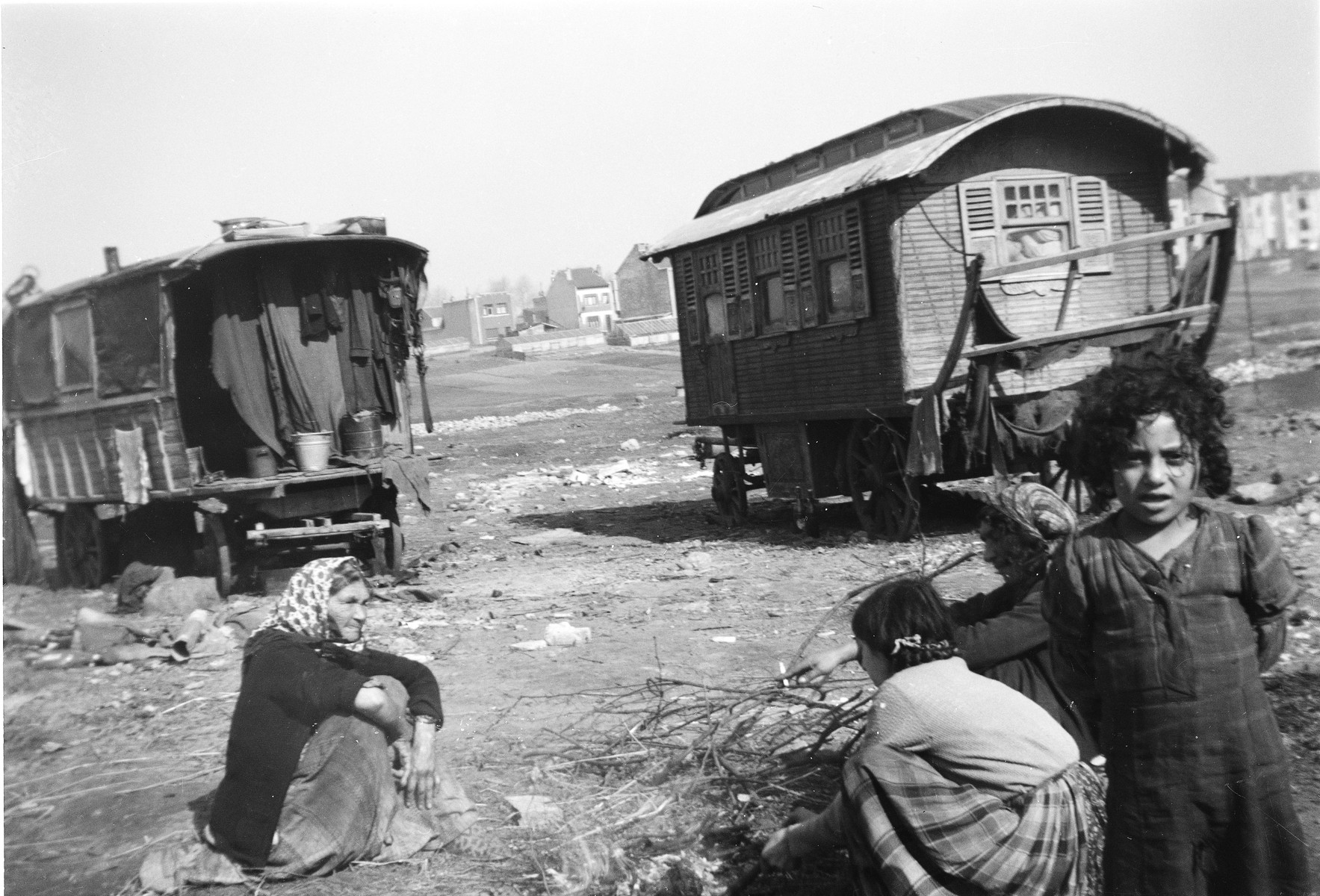 A young Romani girl stands facing the camera, with several seated women and two caravans behind her.