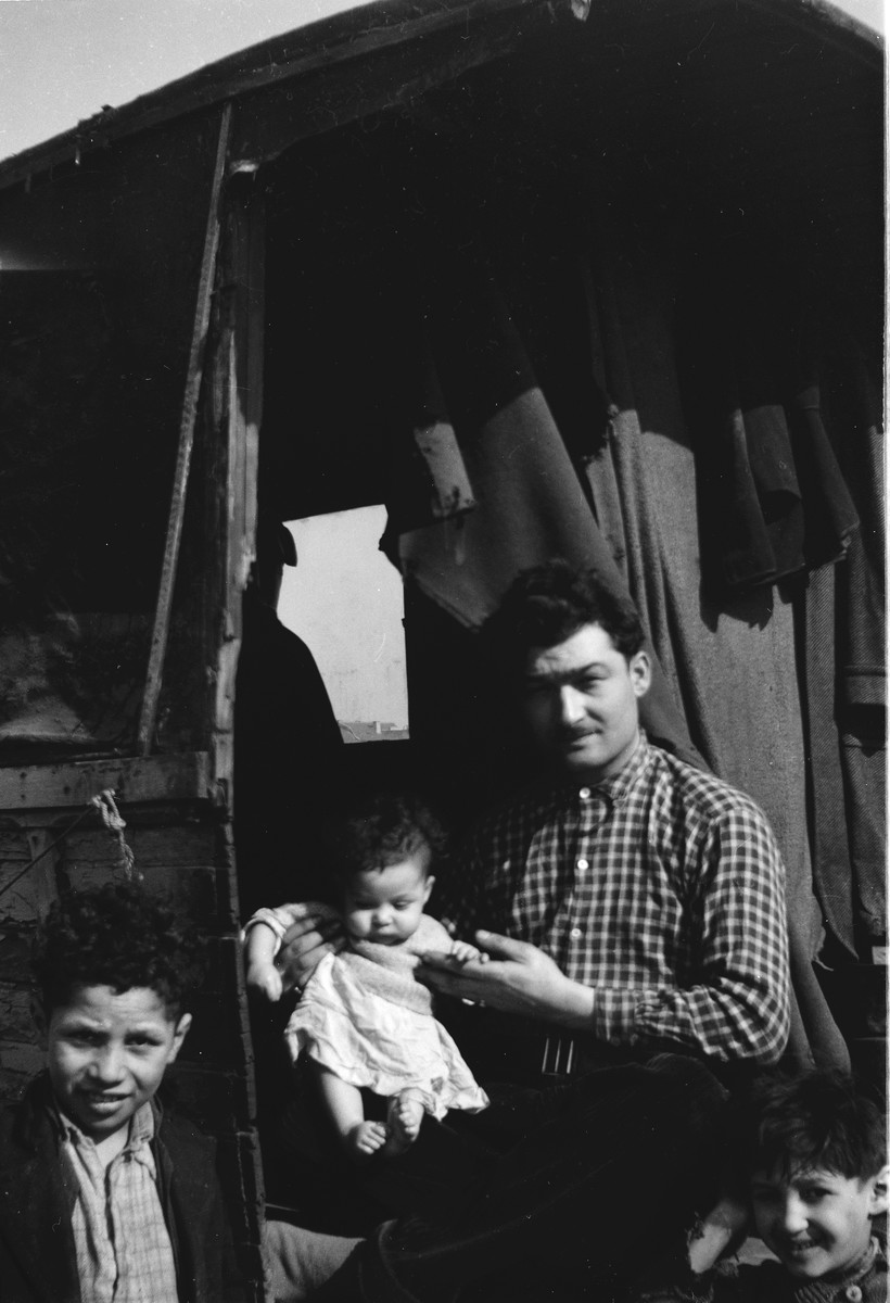 A Romani man holding a baby sits in the doorway of his caravan, with two young boys on either side.