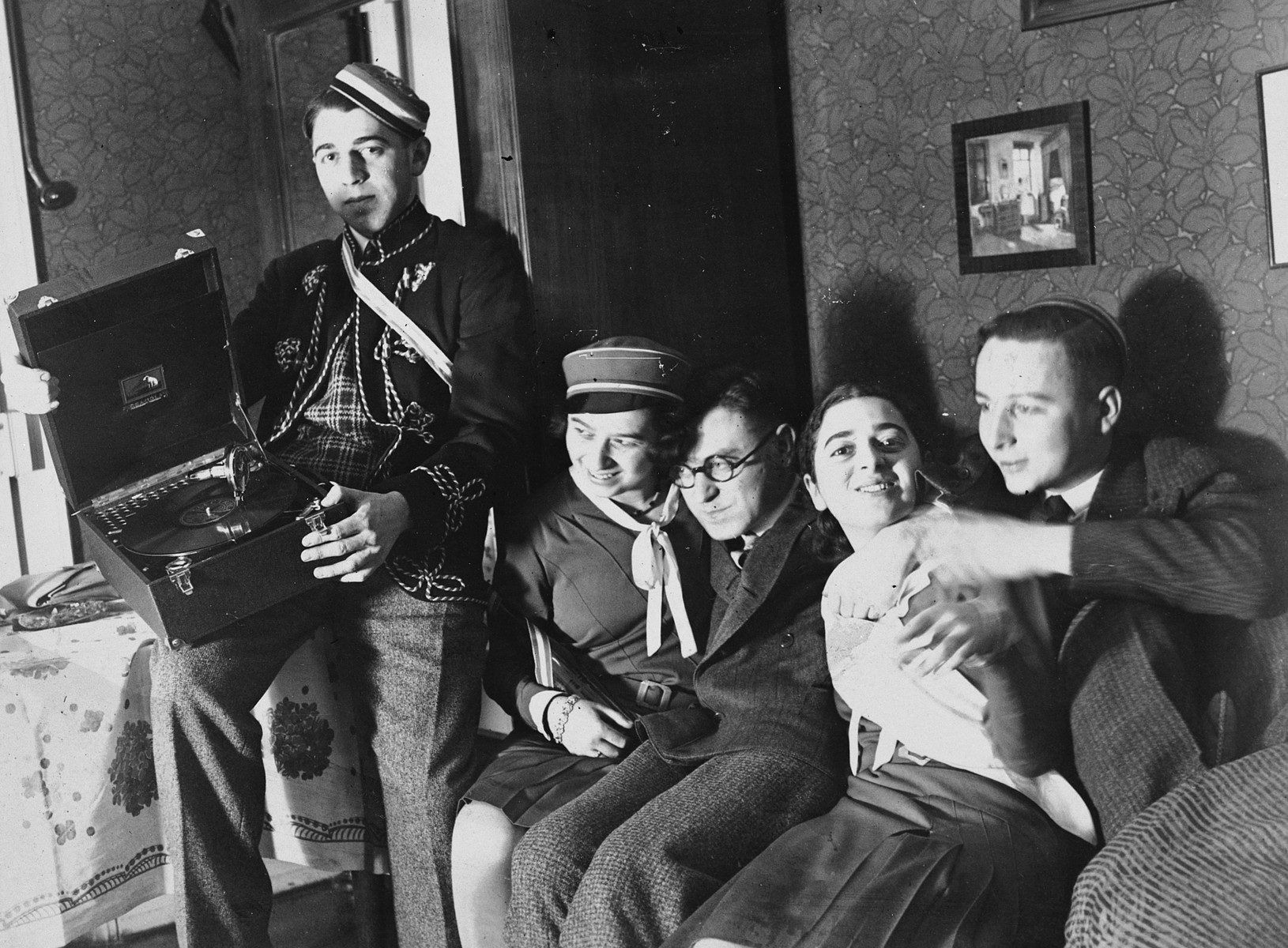 Five German-Jewish university students squeeze together on a couch.  One is holding a phonograph.

From left to right are Heinz Bähr, Ruth Bähr and Margot Bähr is second from the right.  Heinz is wearing the cap and jacket of the KC Jewish fraternity.