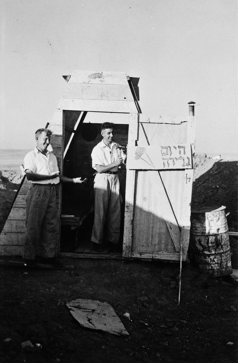 Two kibbutz members stand outside a rustic shack next to a sign advertising "Ice Cream Today."