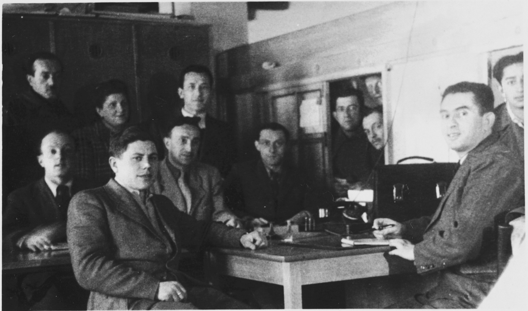 A group of men pose inside an administrative office in the Foehrenwald displaced persons' camp.

Pictured in the bottom left is Sam Spiegel, originally from Kozienice.  Also pictured is Fishl Burakowski, seated behind the desk in the center of the photo. Seated between Sam Spiegel and Fishl Burakowski is Gad Goldman.