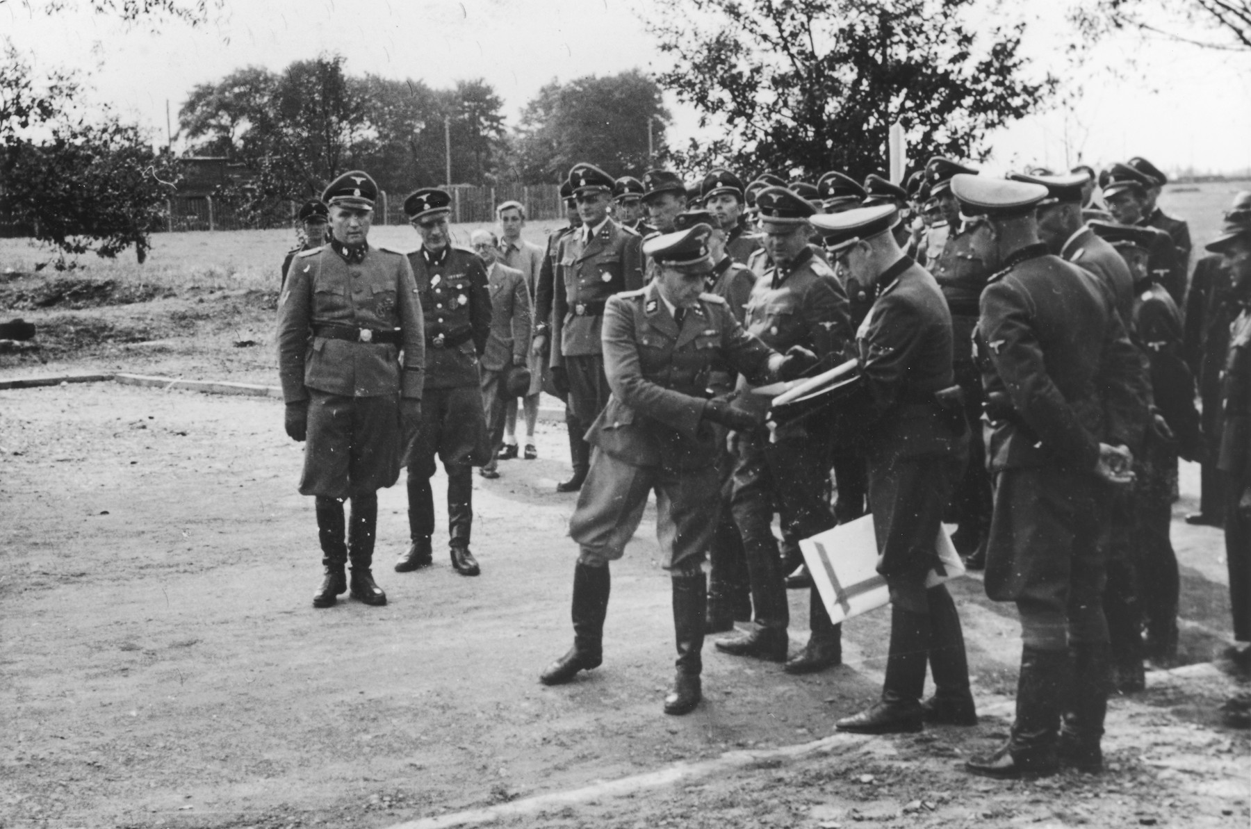 Nazi officers watch the presentation of a document during the dedication of the new SS hospital in Auschwitz.

The original caption reads "Die Ubergabe" (the handover).  The ceremony marks the transfer of documents and authority from the construction department to the camp upon completion of the project.

Commandant Richard Baer stands on the far left. Karl Bischoff, head of construction in Silesia, is receiving the official documents from (most probably Werner Jothann) in preparation for handing them to Baer.  Karl Moeckel is looking down at the exchange of documents.  Also pictured are Drs. Enno Lolling, Eduard Wirths and Carl Clauberg.  Heinz Baumkoetter is in the back looking up slightly.