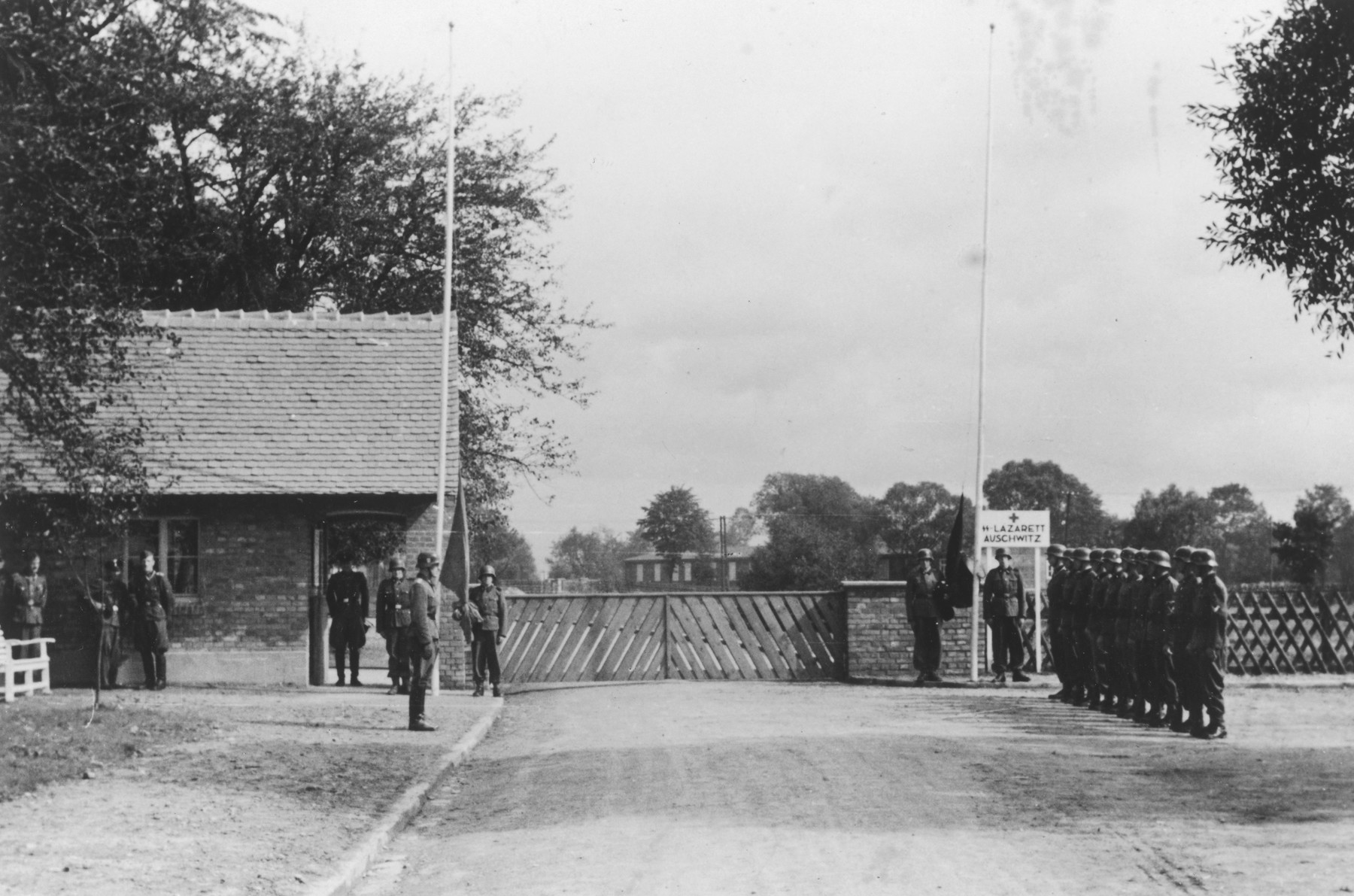 Nazi troops stand at attention during the dedication of the new SS hospital in Auschwitz.  

The official caption reads "Einweihung des SS-Lazarettes in Auschwitz" (Dedication of the SS Hospital in Auschwitz).

Pictured is the entrance to the SS hospital with gate, gate house and flag posts.