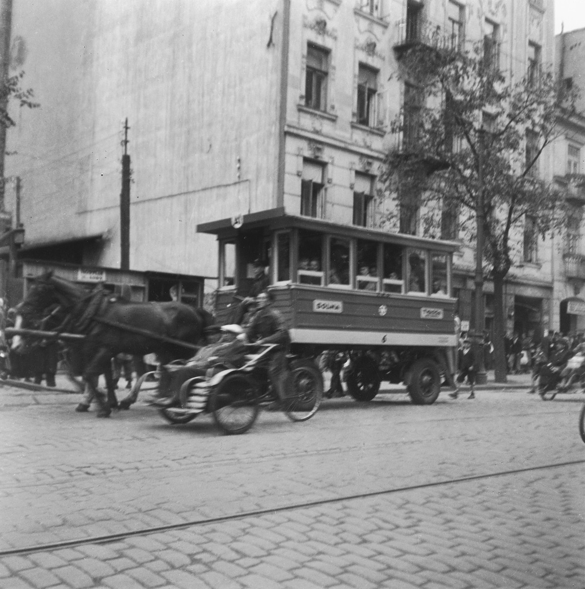 Horse-drawn streetcars and rickshaws ride down a street in the Warsaw ghetto. 

Joest's original caption reads: "There were horse-drawn streetcars, and there were the rickshaws.  They were faster, but they were almost more expensive, I was told."