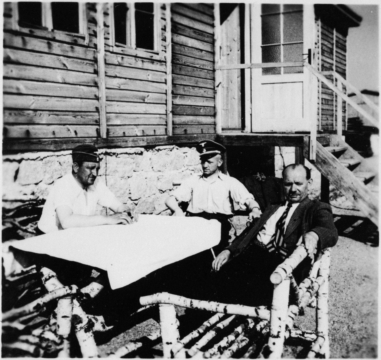 SS officers and an unidentified man sit at a picnic table outside a building at the Gross-Rosen concentration camp.

Pictured on the left is the commandant, Arthur Roedel.  His adjutant, Kuno Schramm is in the center.