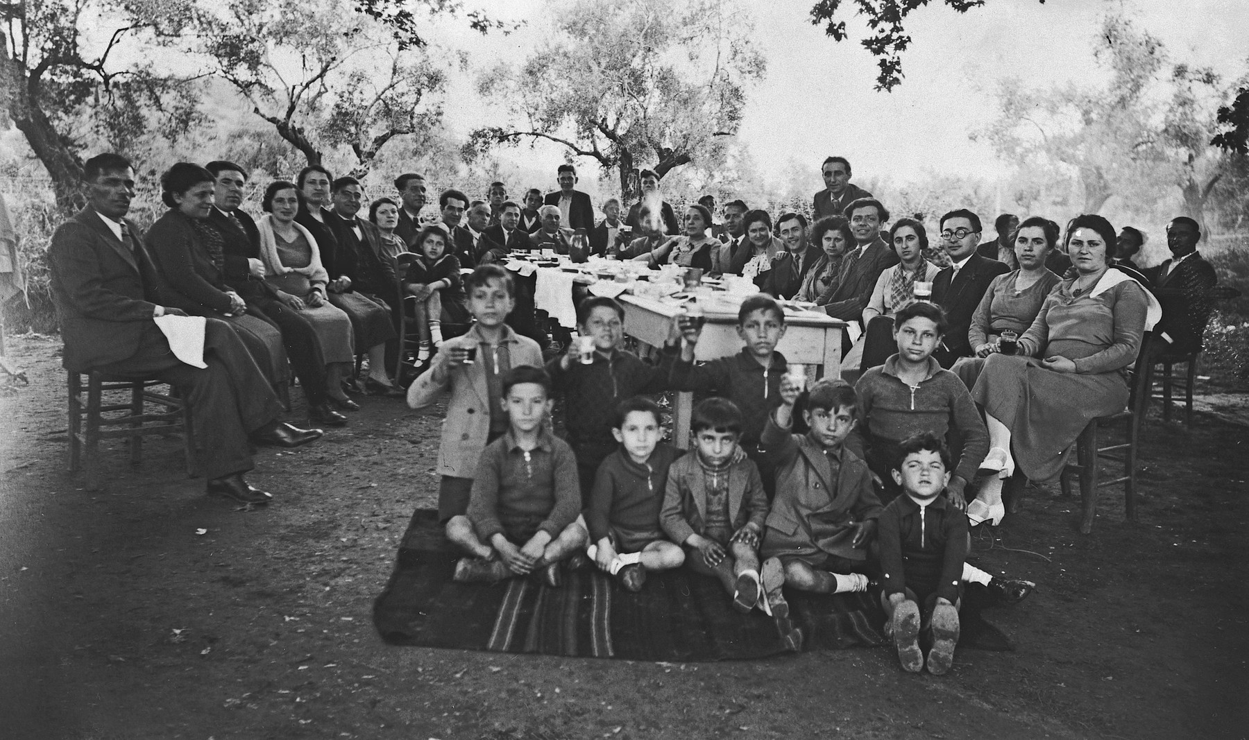Employees and family of the National Bank of Greece in Arta gather for a group portrait during an outdoor celebration.

Michael Matsas is seated in the front, second from the left.  His parents, Leon and Esther Matsas, are seated behind him, third and fourth from the left.