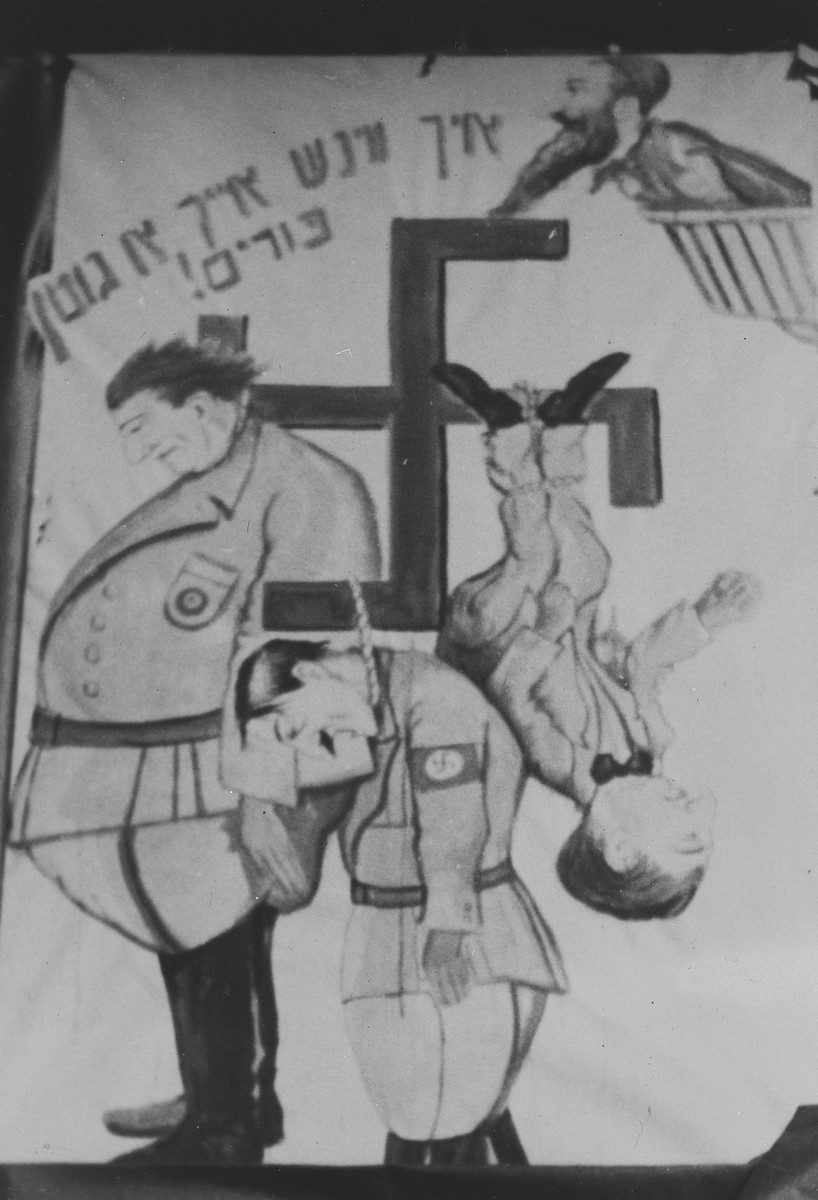 Photograph of a poster in the Landsberg DP camp with a cartoon illustration of Hitler, Goering and possibly Goebbels hanging from a swastika.

Tha caption reads, "I wish you a good Purim."