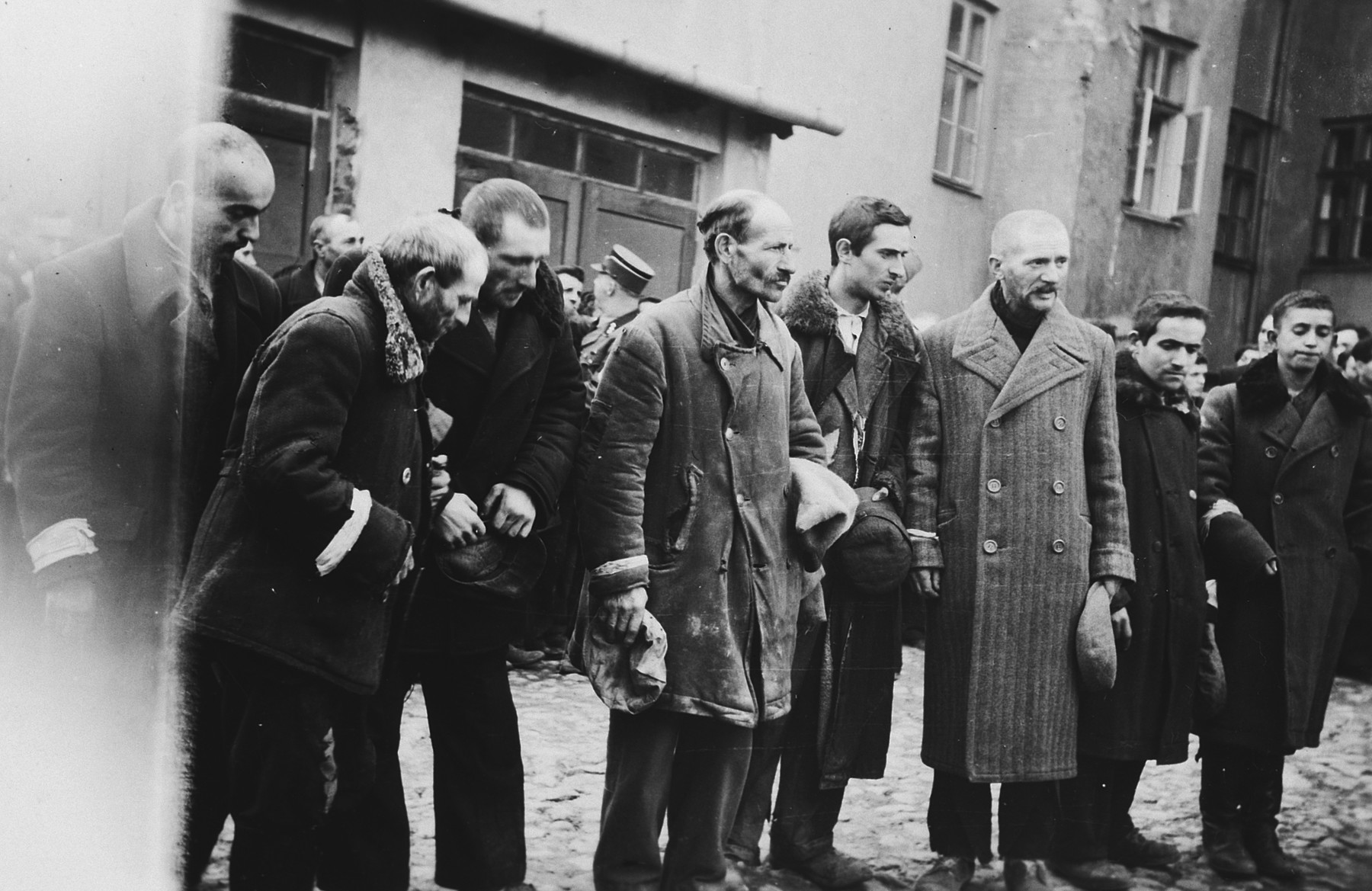 Jewish men are forced to stand in a line holding their hats in their hands [perhaps right after having had their beards forcibly shaven].
