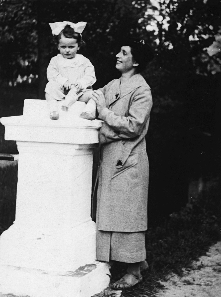 A young Polish-Jewish toddler with a large bow sits on top of a pillar supported by her mother.

Pictured are Sala and Bela Perec.