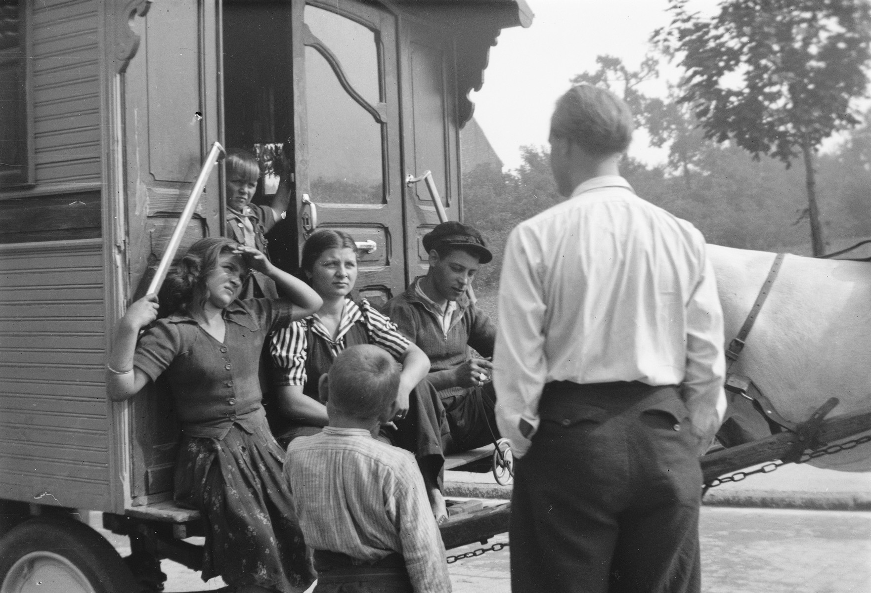 A group of Romani children gathered around the entrance to their caravan converse with with their friend, Jan Yoors (with back to the camera).

The caption in "The Heroic Present" reads, "Jan Yoors (back to camera) and Lovara friends, Europe, 1930s."