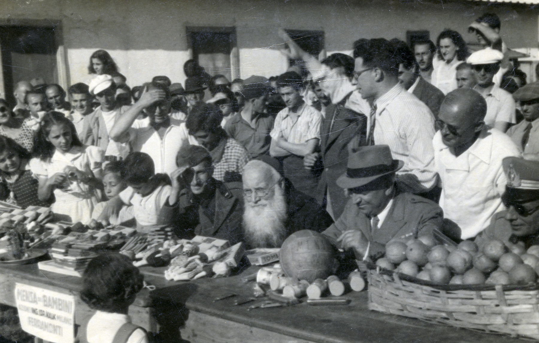 Internees at Ferramonti gather by a table displaying various goods.