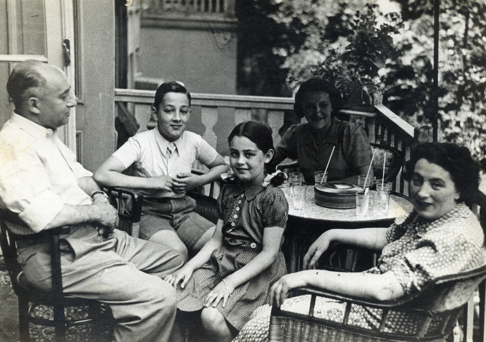 Isaak Krzywanowski sits on a porch with his wife and children.

From left to right are Isaak, Jacob, Rebecca and Sara Krzywanowsk.  Seated in the background is Isaak's sister, Bronia Krzywanowski.  She was born in Plonsk, Poland in 1902 and  survived the war in hiding in Amsterdam. In 1949, she immigrated to Israel, and settled in Tel Aviv.
