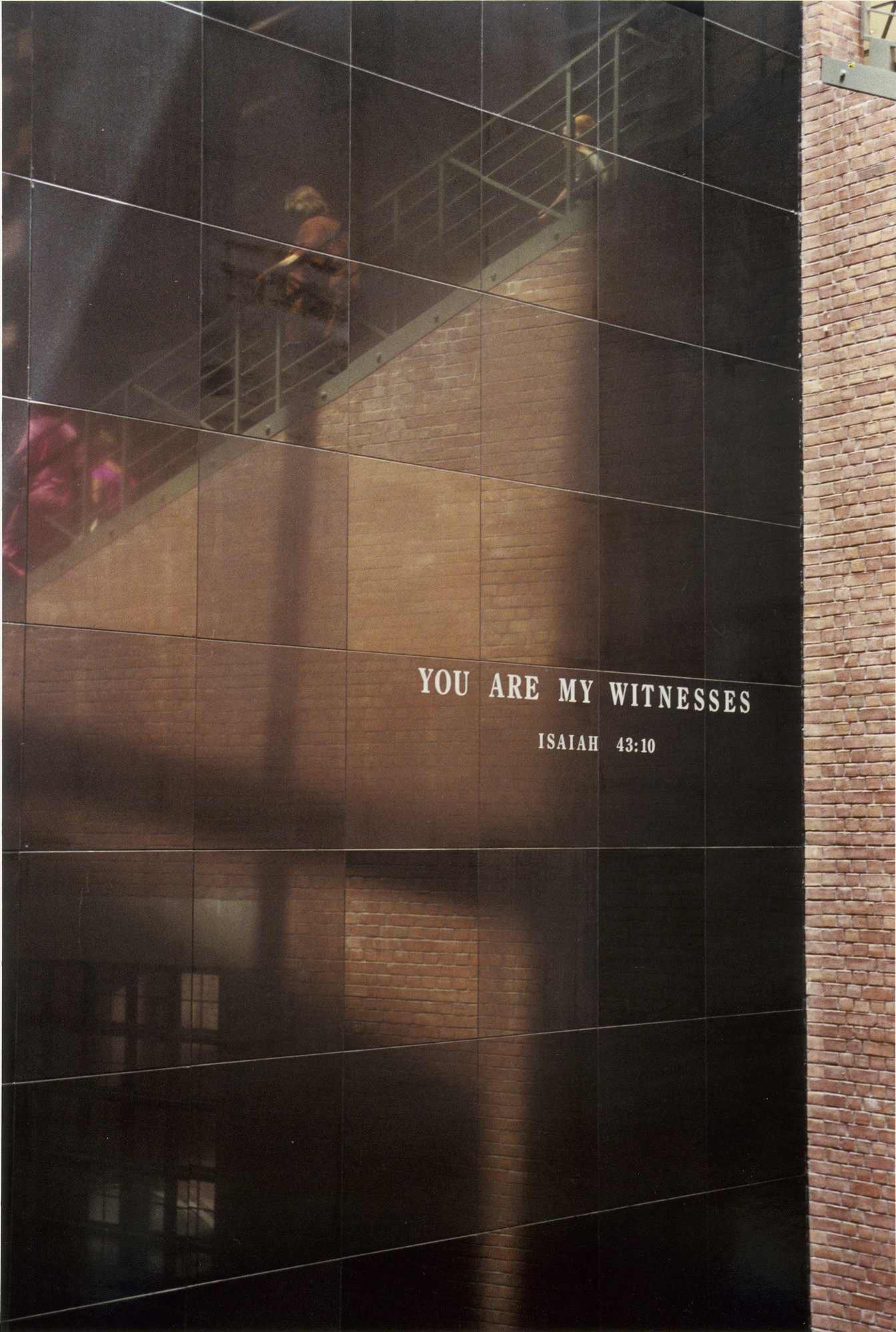 Black granite wall bearing the quote "You are my Witnesses" located in the Hall of Witness at the U.S. Holocaust Memorial Museum.

Reflected on the wall is the grand staircase with visitors.