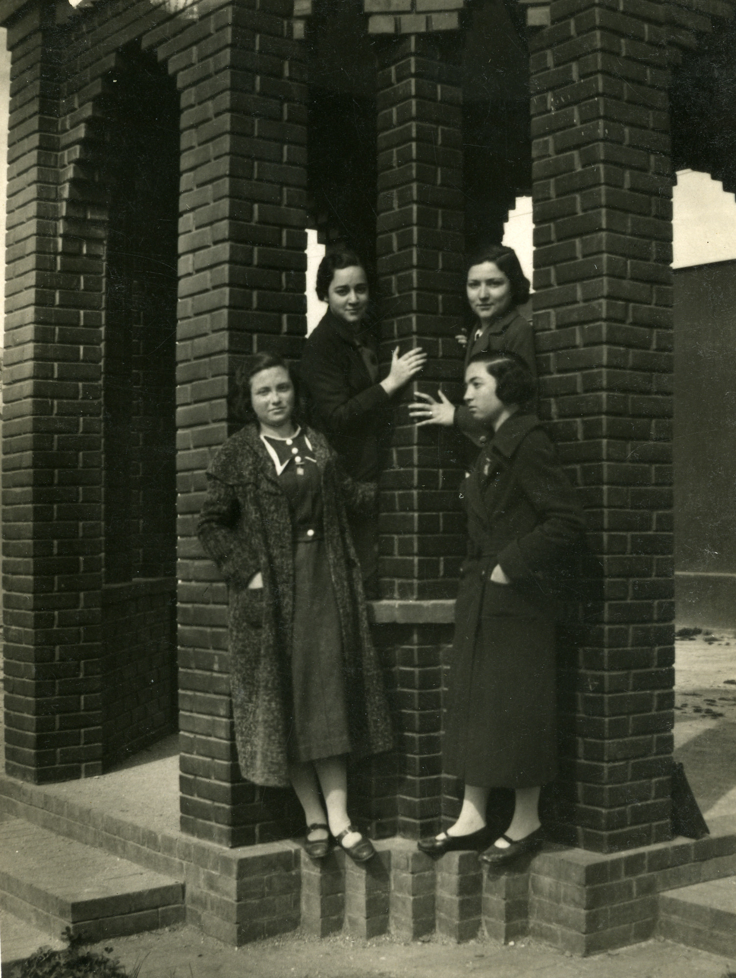 Four young women stand on the ledge of a brick structure under the bridge, in Salonika in 1940.

Jenny Noam (nee Ezratty) is standing in the front row on the left.
