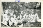 A group of toddlers sit on the grass at a birthday party.