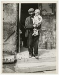 A man holds his young child in the entrance to a building in a displaced persons camp in Austria

Handwritten caption on the back of the photograph reads: "In a Displaced persons Camp in Austria."