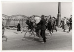 Pedestrians cross a bridge during the siege of Warsaw; one Polish man in the center carries bedding on a bicycle.