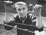 Aleksander Kulisiewicz poses holding on to the barbed wire fence at a former concentration camp [possibly Sachsenhausen] during a performance of his collected concentration camp music.